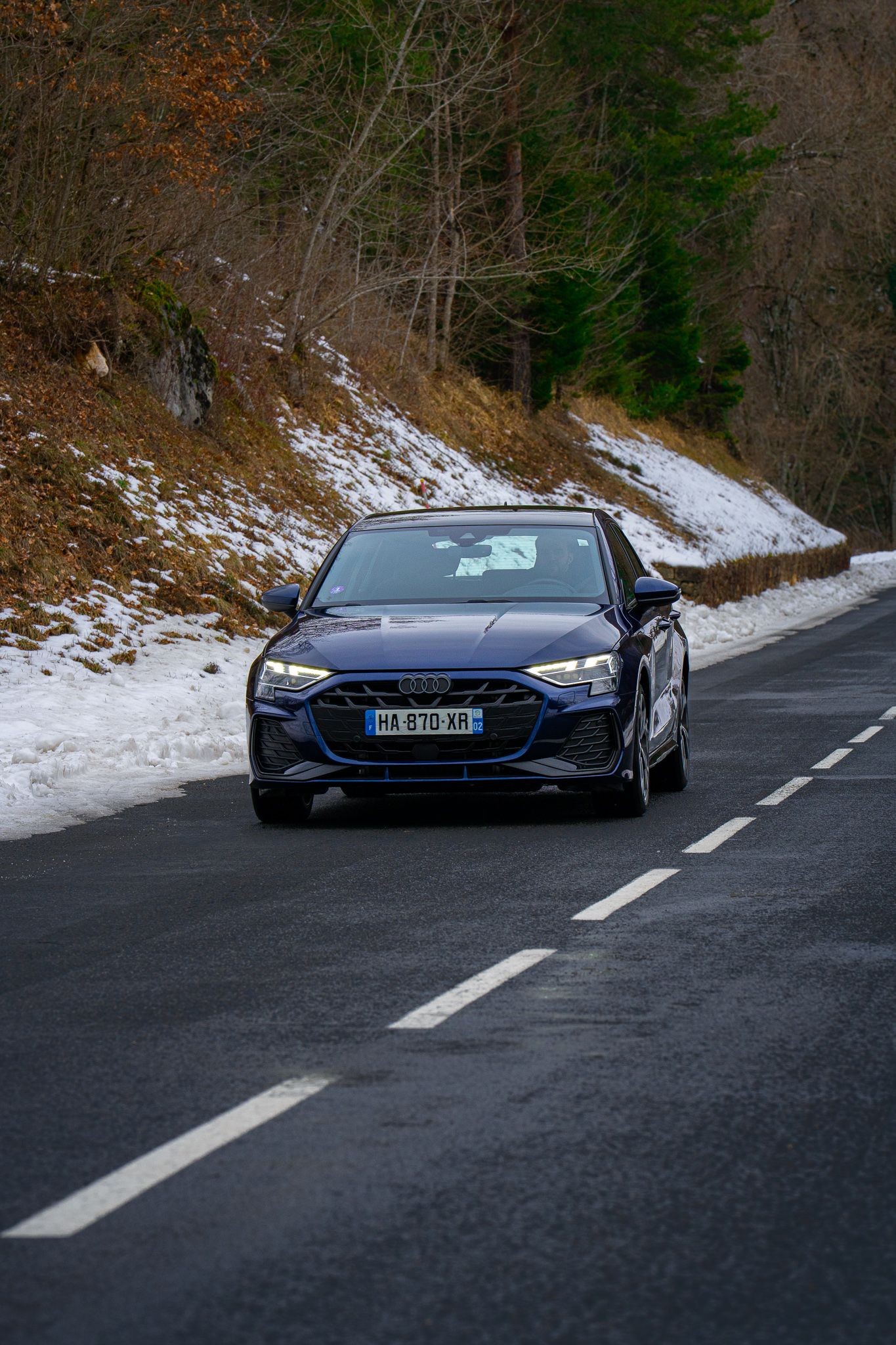 A sleek blue sports car navigates a winding mountain road with patches of snow visible on the roadside.
