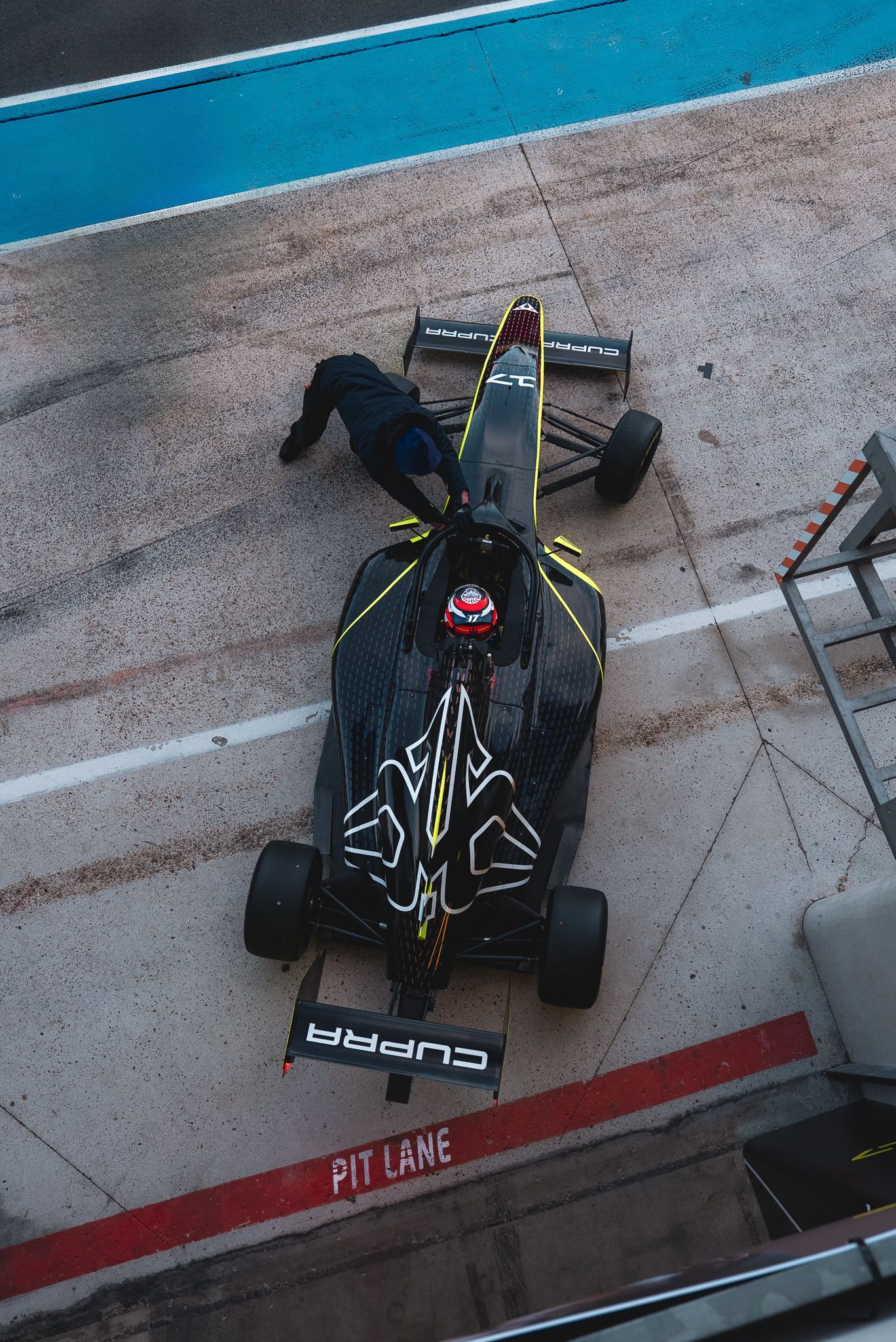 High angle view of a black Formula 1 race car with yellow accents parked in a pit lane during testing or practice session.