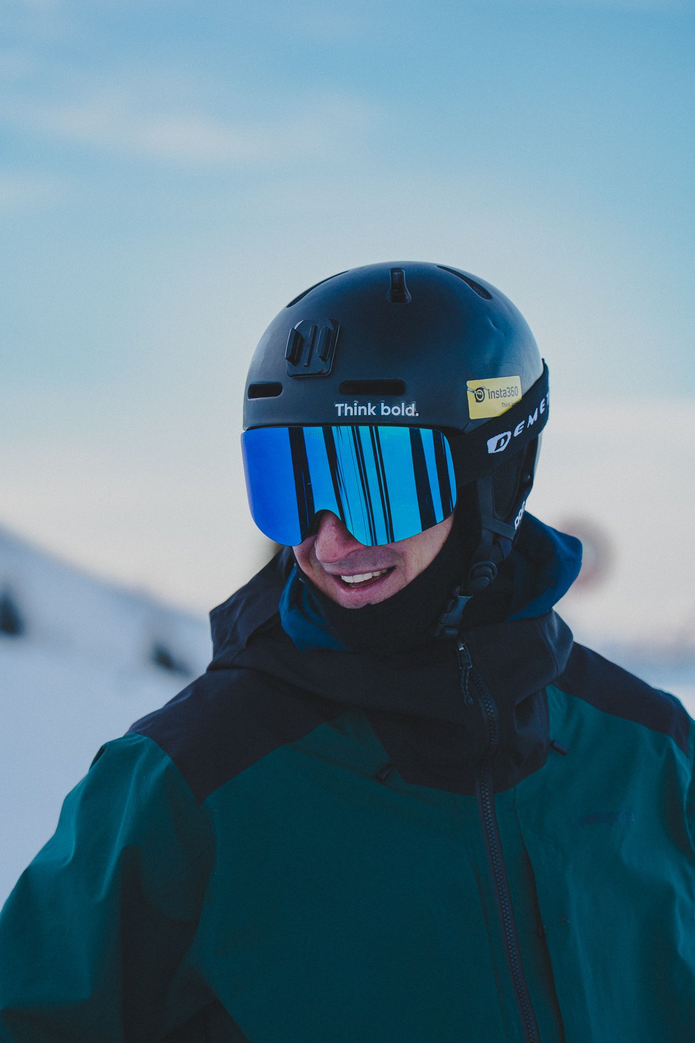 Skier wearing black helmet with blue mirrored visor and dark winter gear against snowy mountain backdrop.