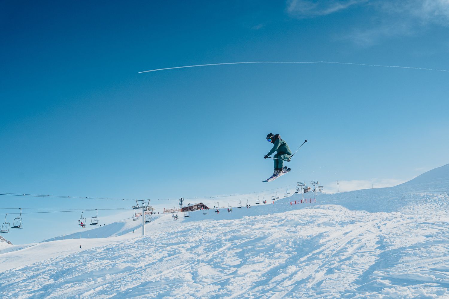 A skier jumps high in the air against a bright blue sky at a snowy ski resort with chairlifts in the background.
