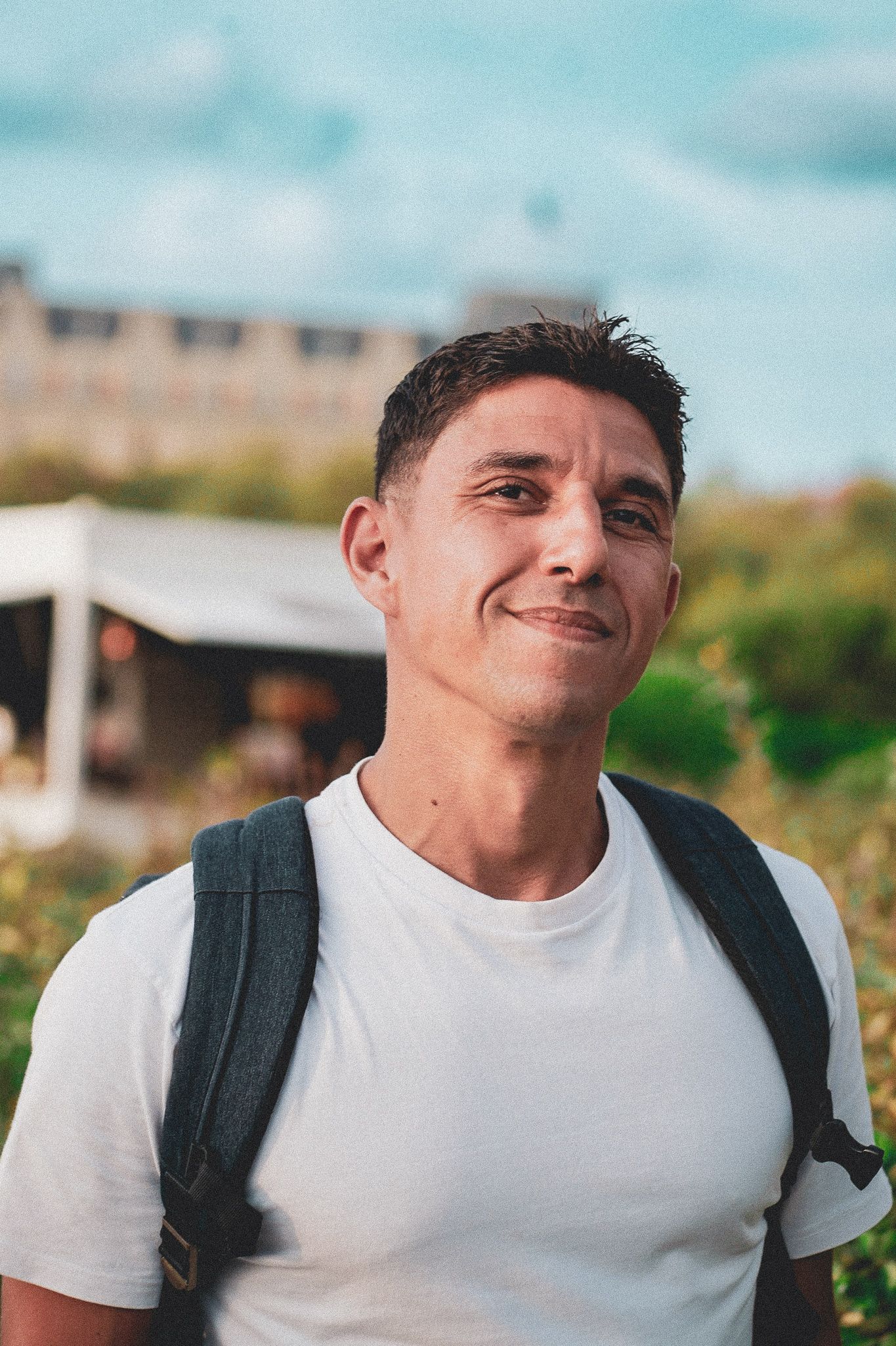 Young person wearing a white t-shirt and backpack smiles at the camera on a sunny day outdoors.