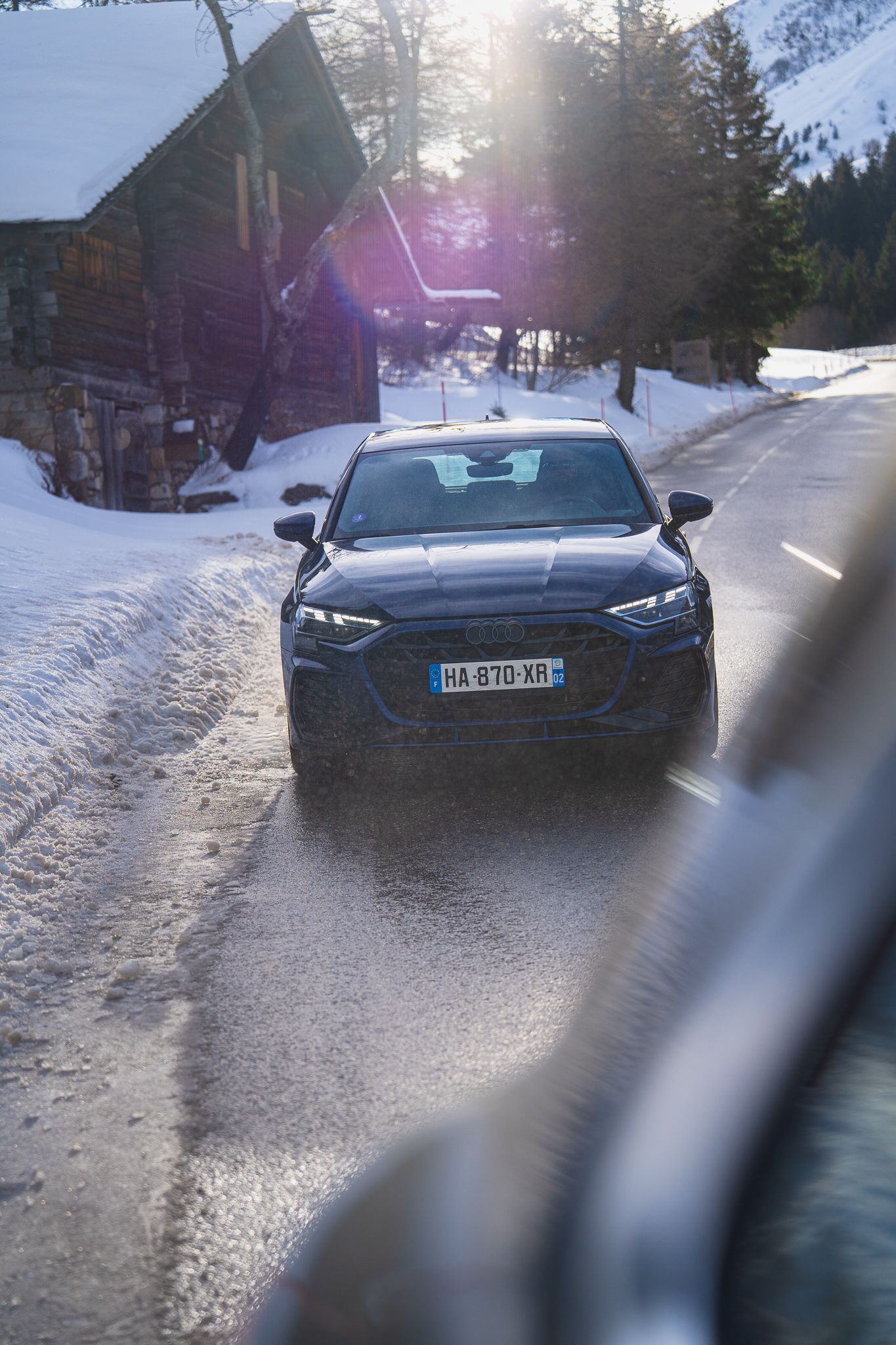A dark BMW car parked on a snowy mountain road with wooden chalets and sunlight peeking through the trees.