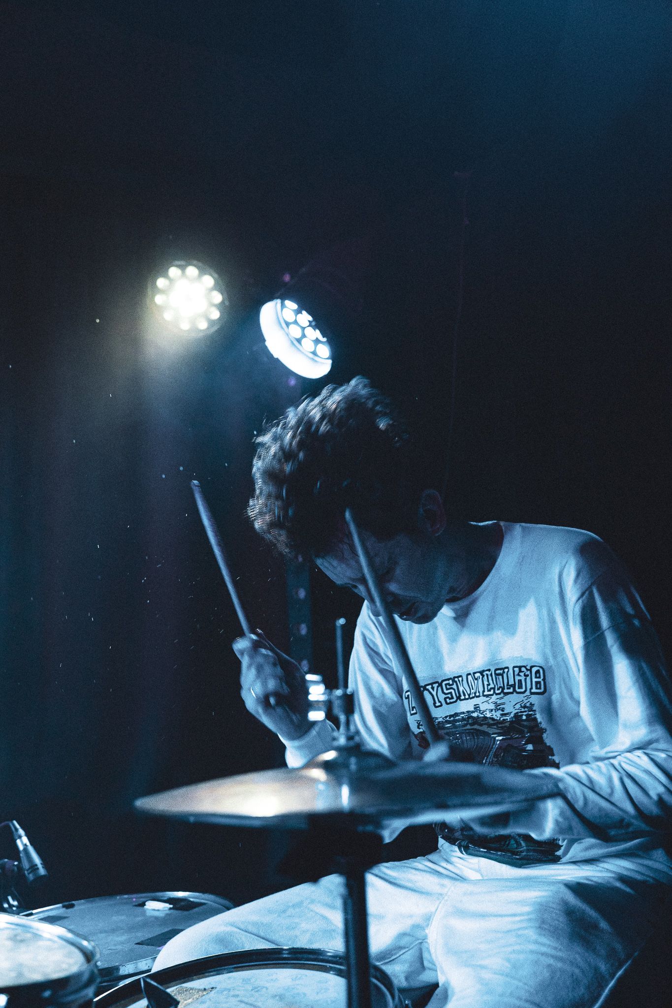 A drummer performs on stage illuminated by bright spotlights in a moody blue atmosphere.