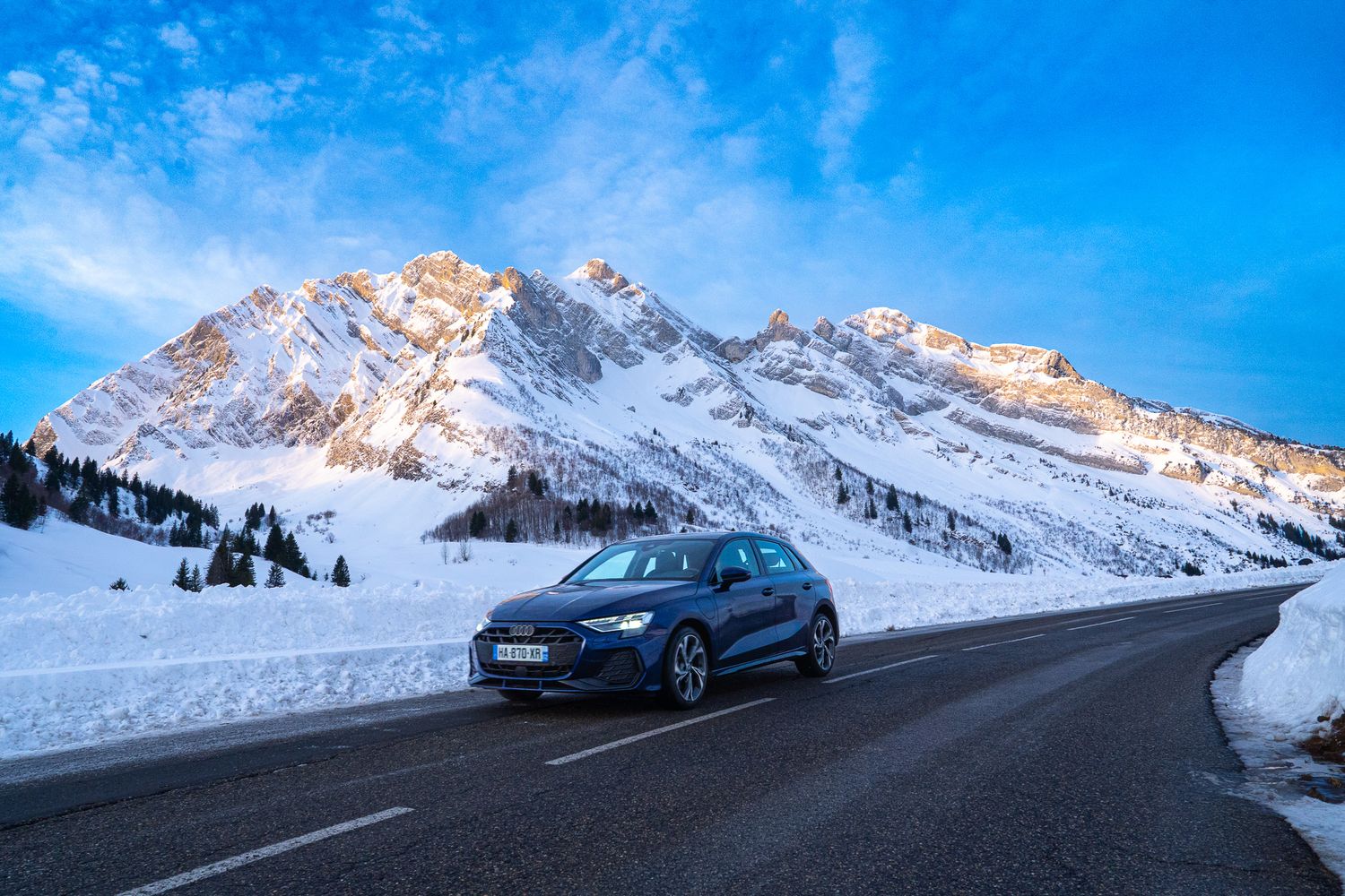 A sleek BMW car drives on a snowy mountain road with snow-capped peaks and blue sky in the background.