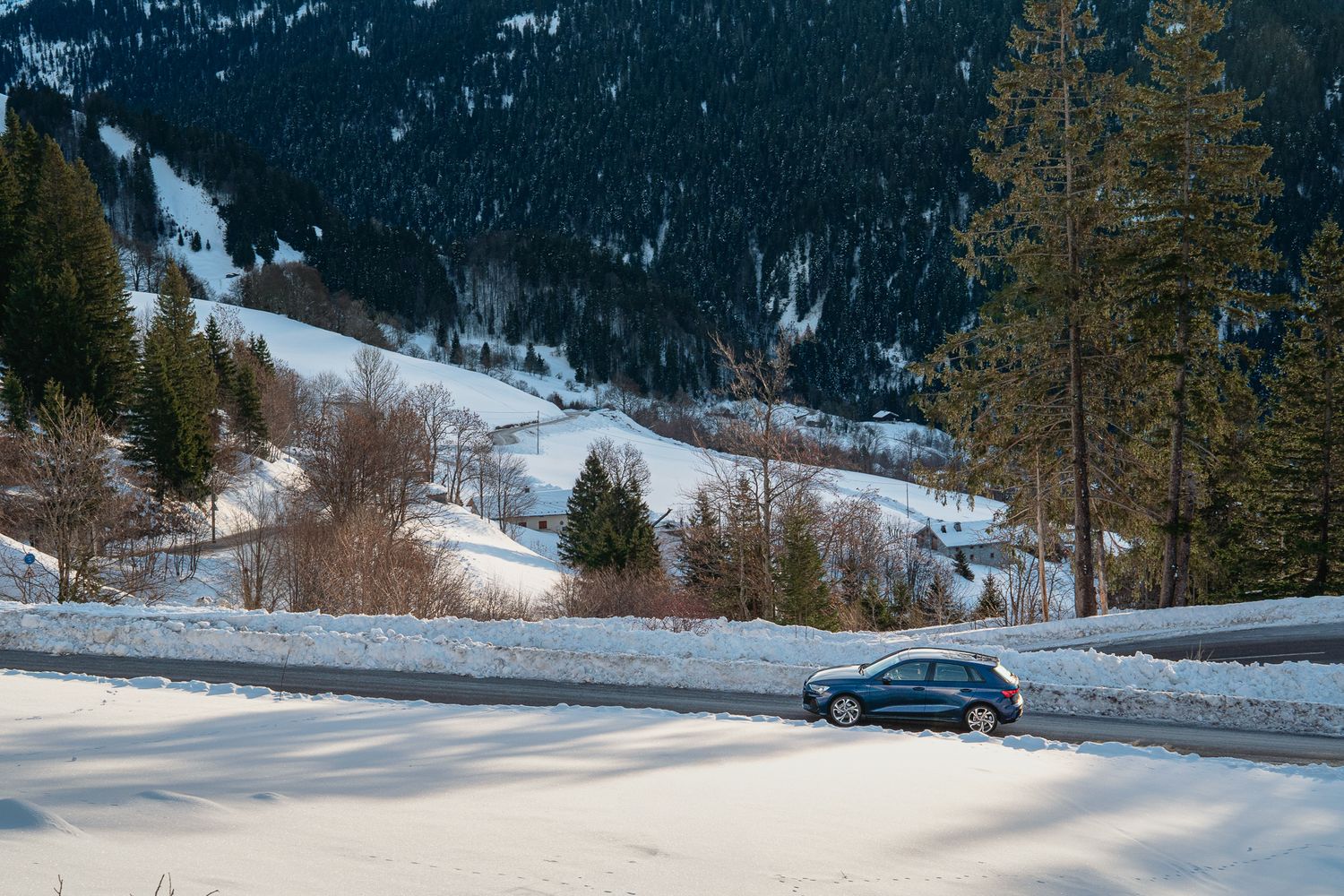 A black car drives along a snowy mountain road with snow-covered evergreen trees and peaks in the background.