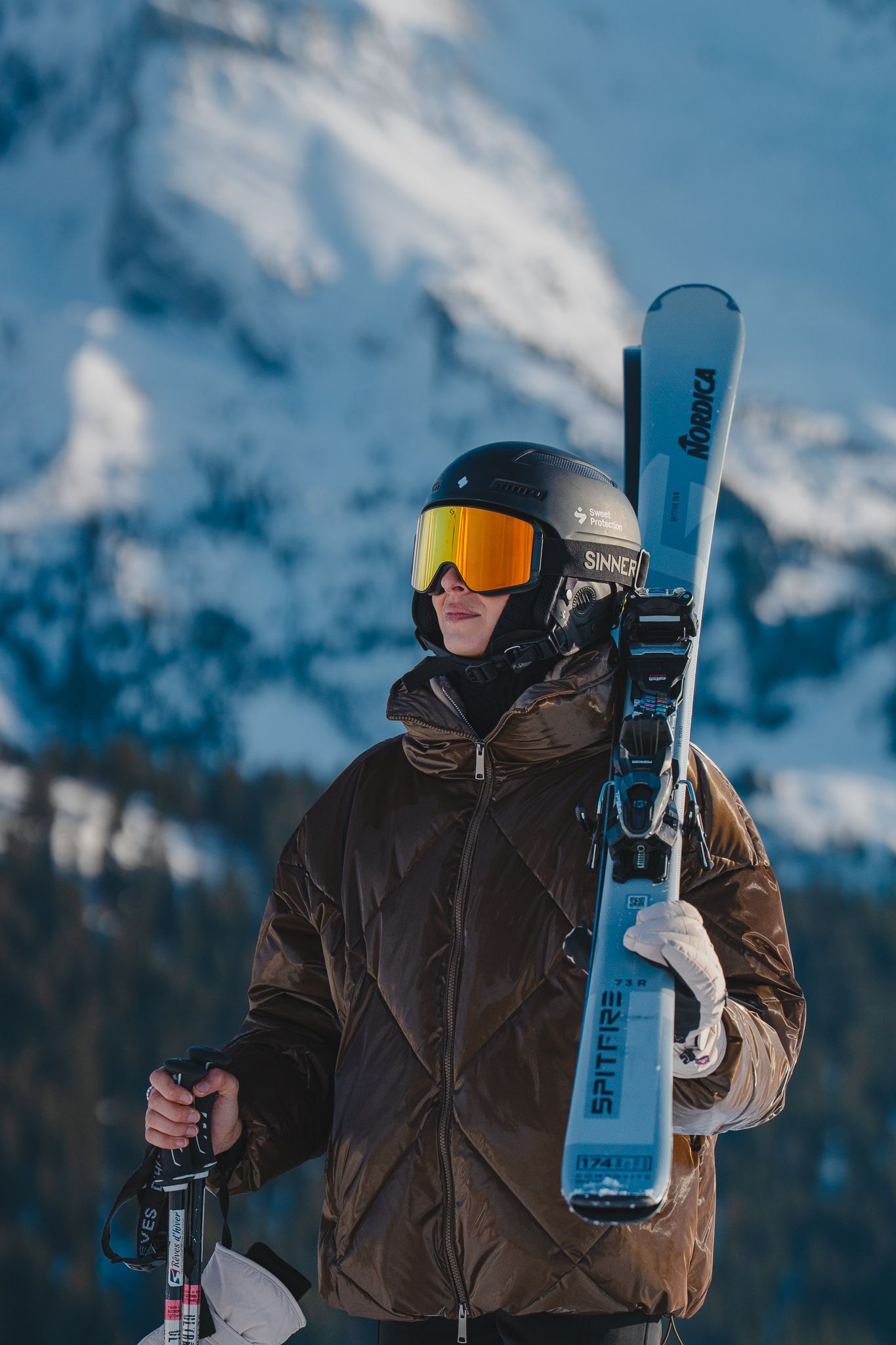 A skier in a brown jacket carries blue skis against a snowy mountain backdrop on a clear winter day.
