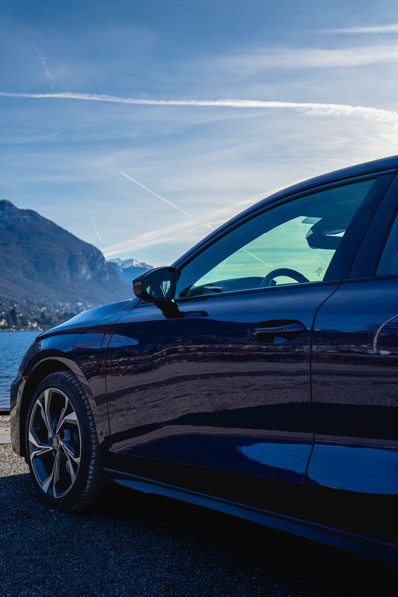 A sleek dark blue car's side profile is showcased against a mountainous backdrop with a bright blue sky.