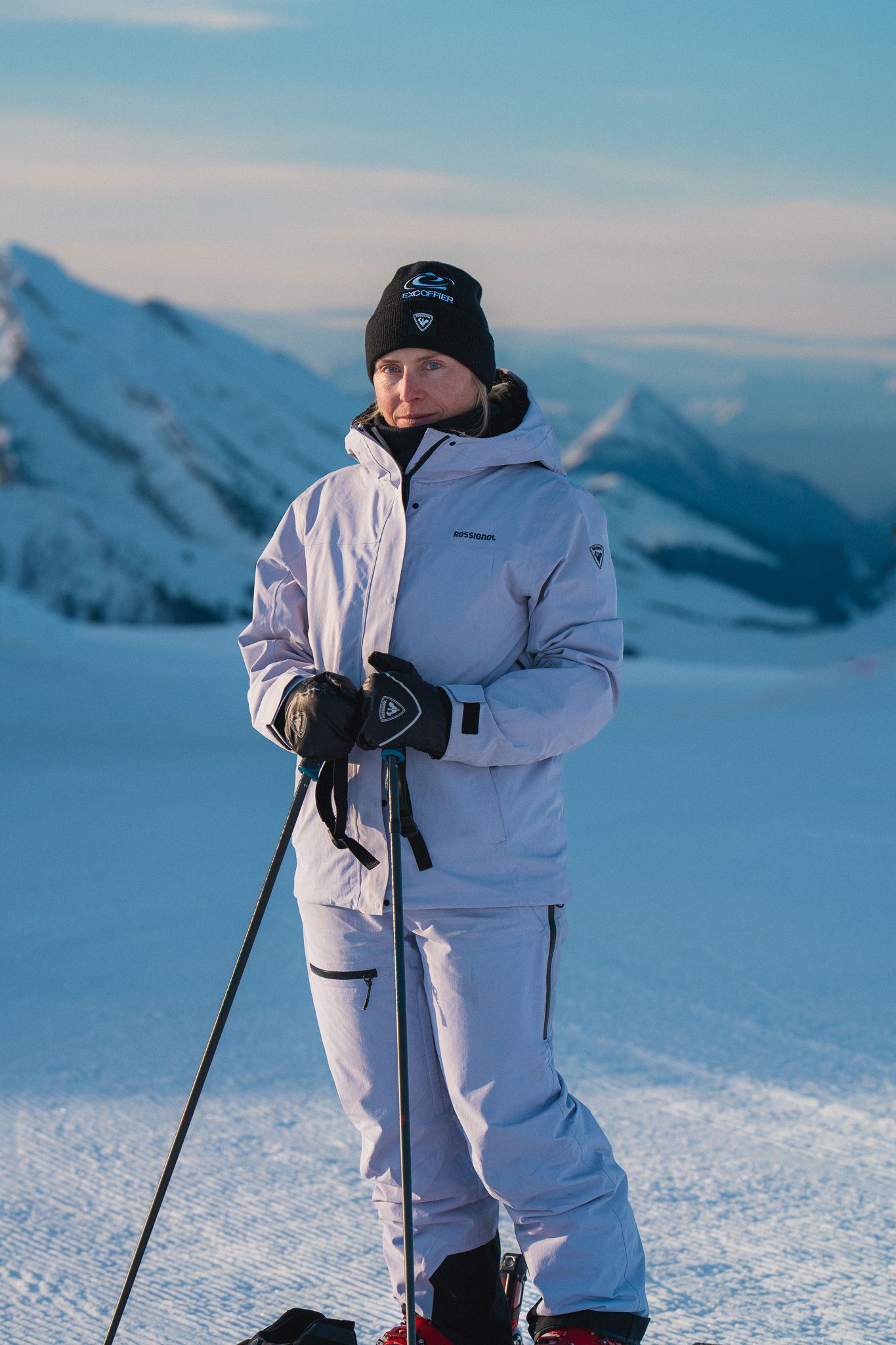A skier in an all-white snowsuit and black beanie stands on snowy slopes with mountain peaks in the background.