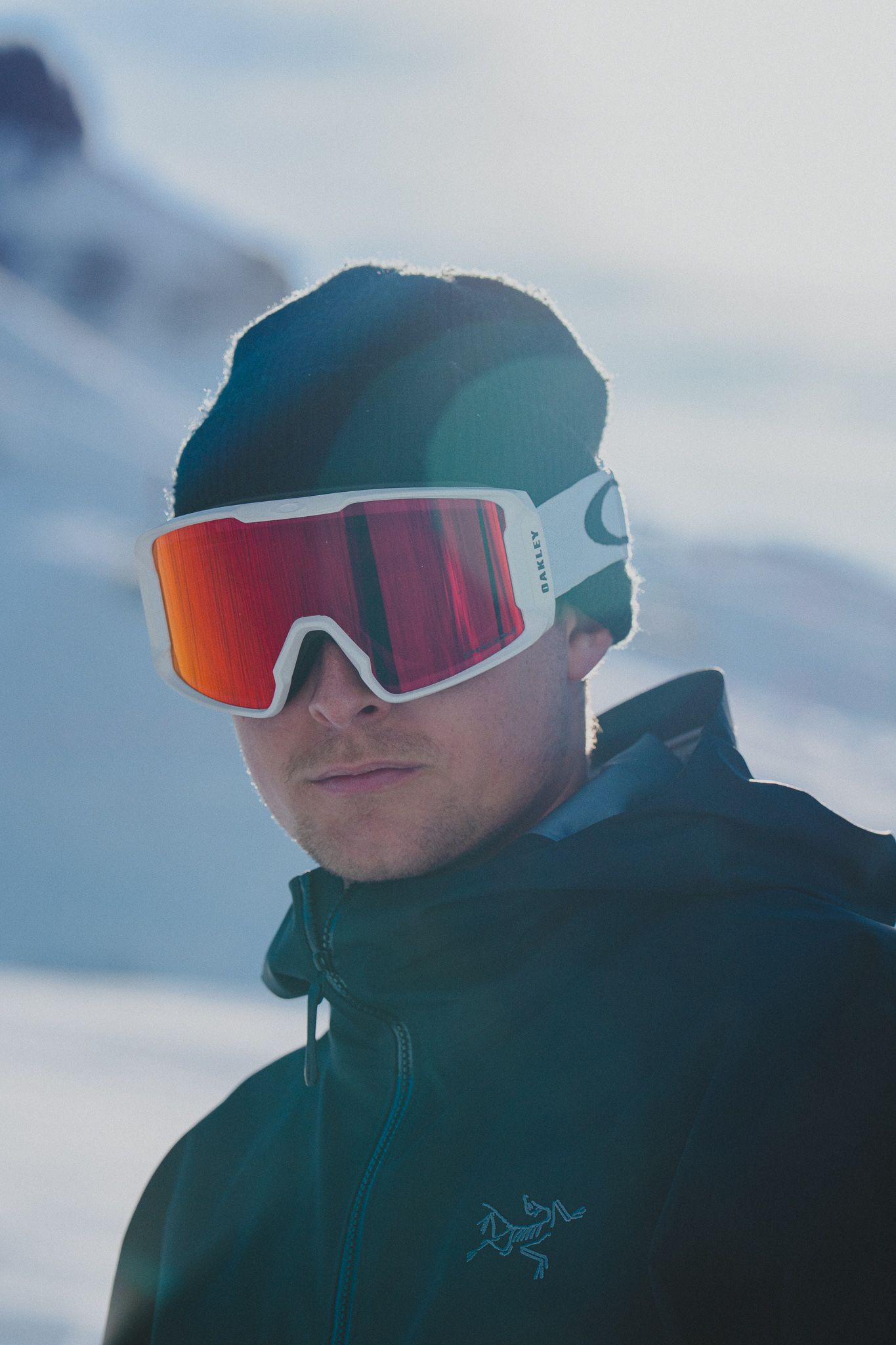 A person wearing reflective ski goggles and winter gear stands against a snowy mountain backdrop.