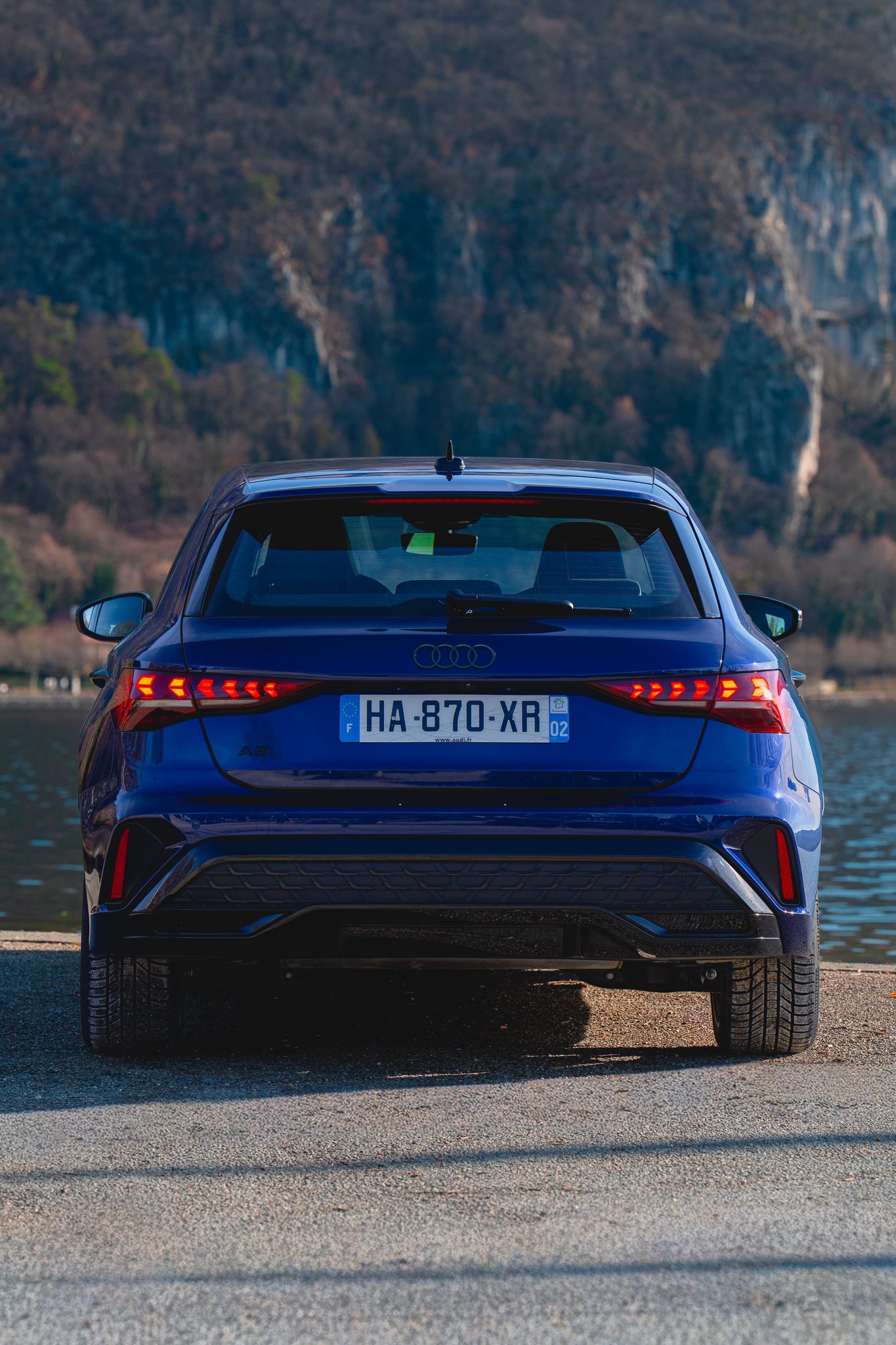 Rear view of a sleek blue Peugeot 408 sports car parked by a lakeside with mountains in the background.
