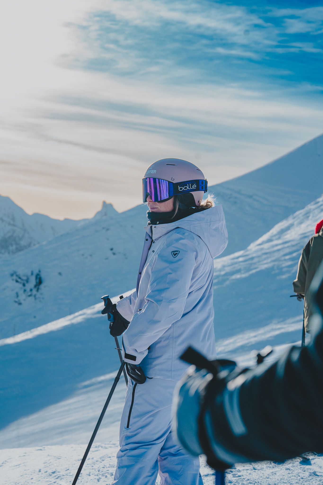 A skier in white winter gear stands against a snowy mountain backdrop with blue sky overhead.