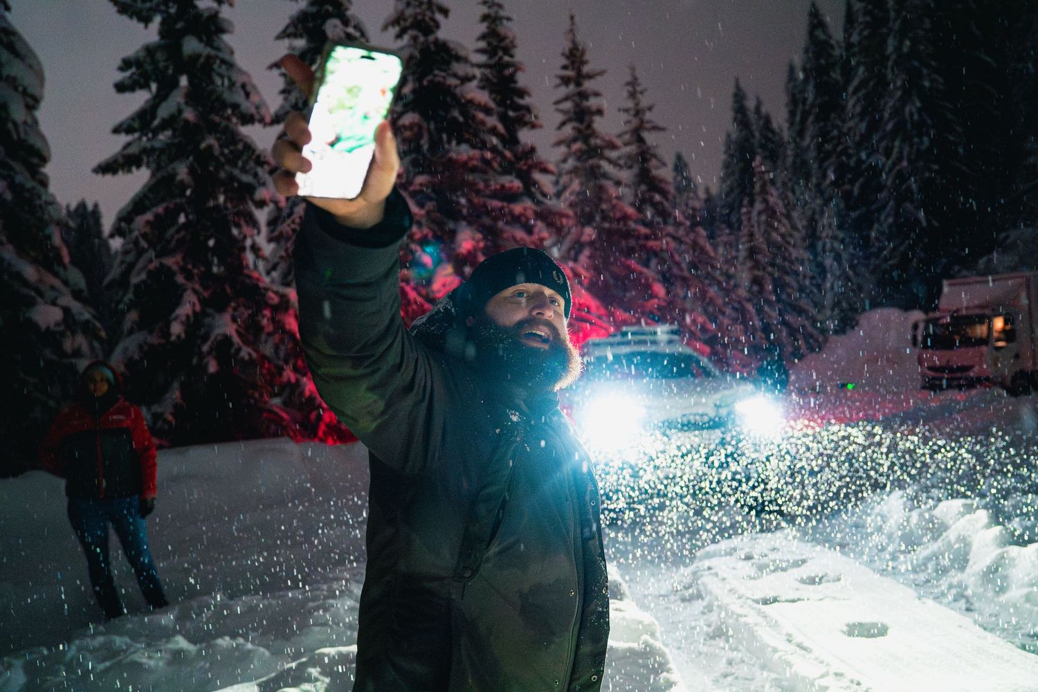 Someone holds up a bright phone light while standing in a snowy forest at night.