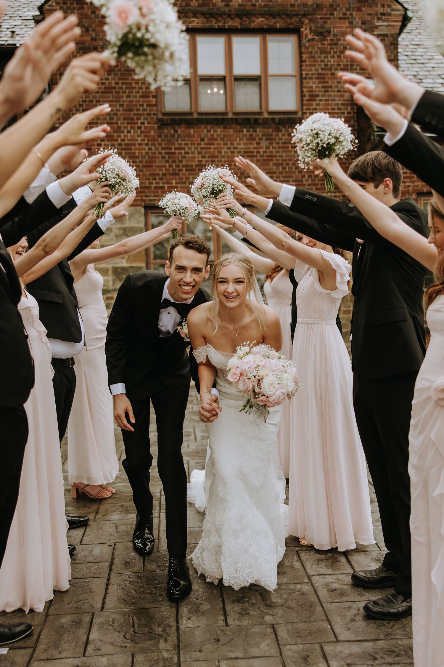 Wedding party celebrating with raised bouquets forming an arch on brick building backdrop.