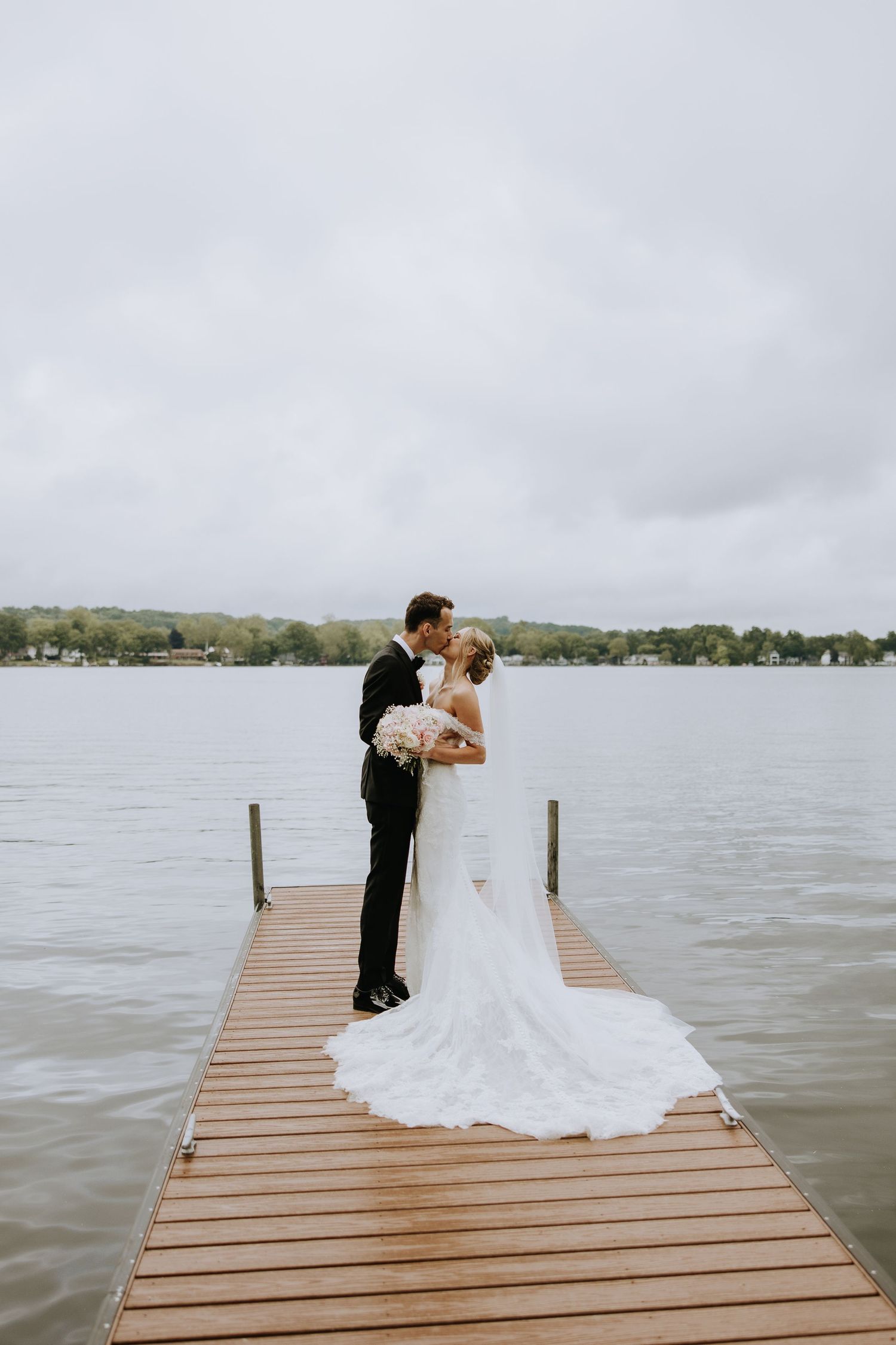 Couple in wedding attire share a romantic moment on a wooden dock overlooking a serene lake.