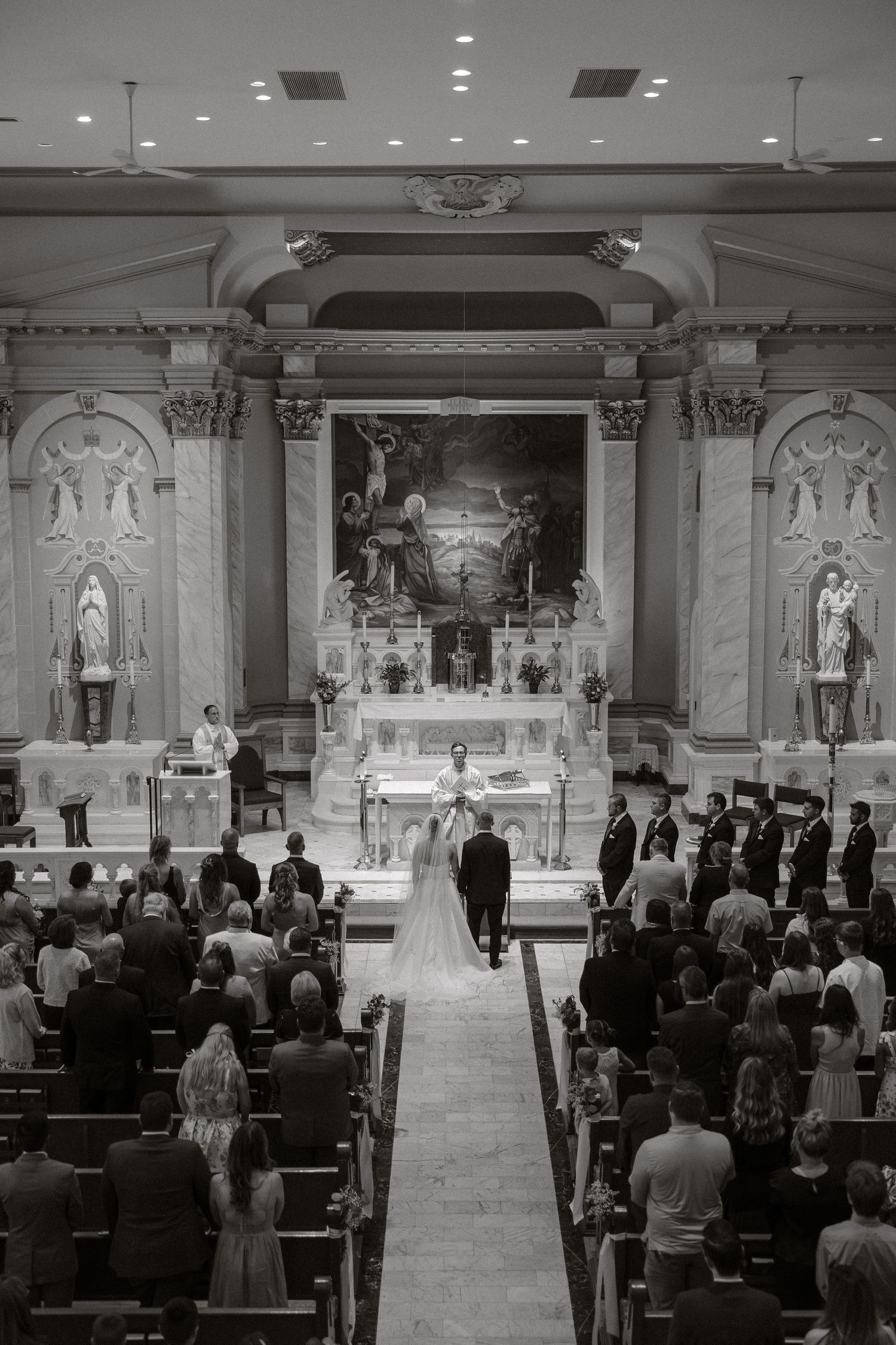 A traditional Catholic church wedding ceremony with guests seated in ornate marble sanctuary and white floral decorations.