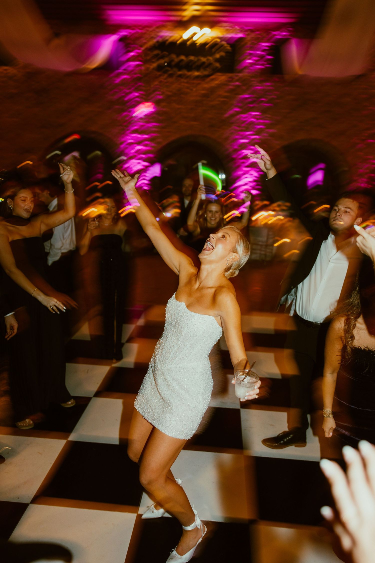 Wedding reception dance party with colorful pink lighting and guests dancing on a checkered floor.
