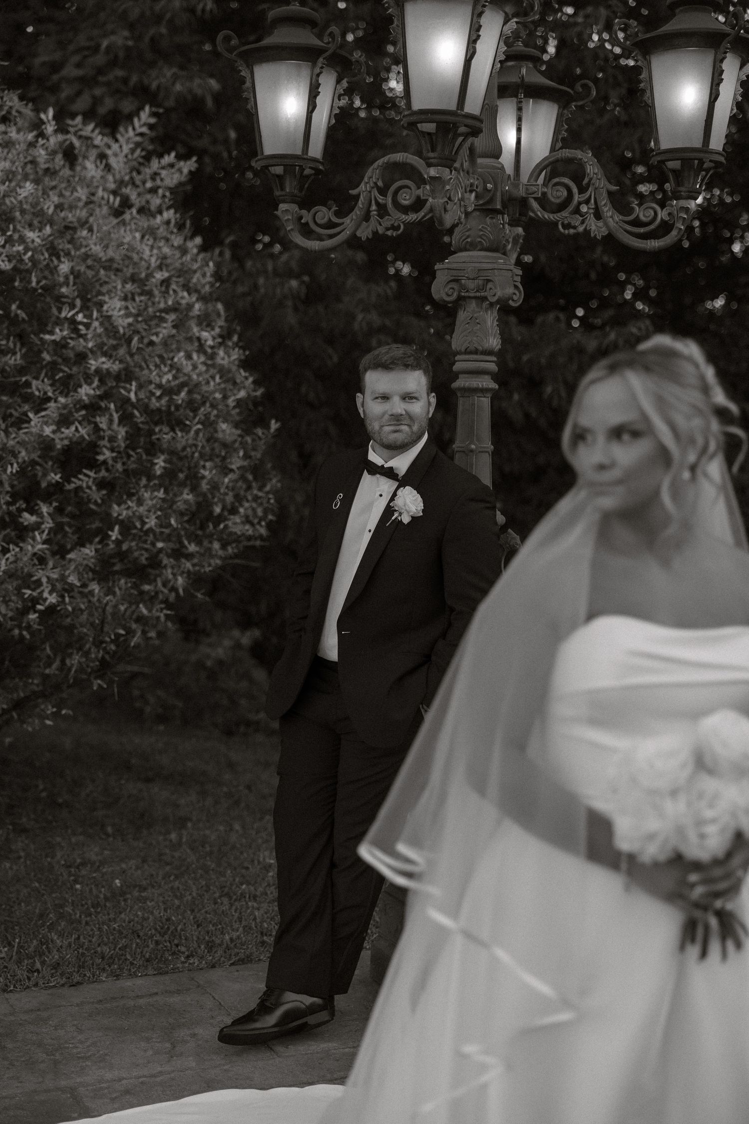 A wedding couple poses for romantic evening photos under ornate lamp posts in a garden setting.