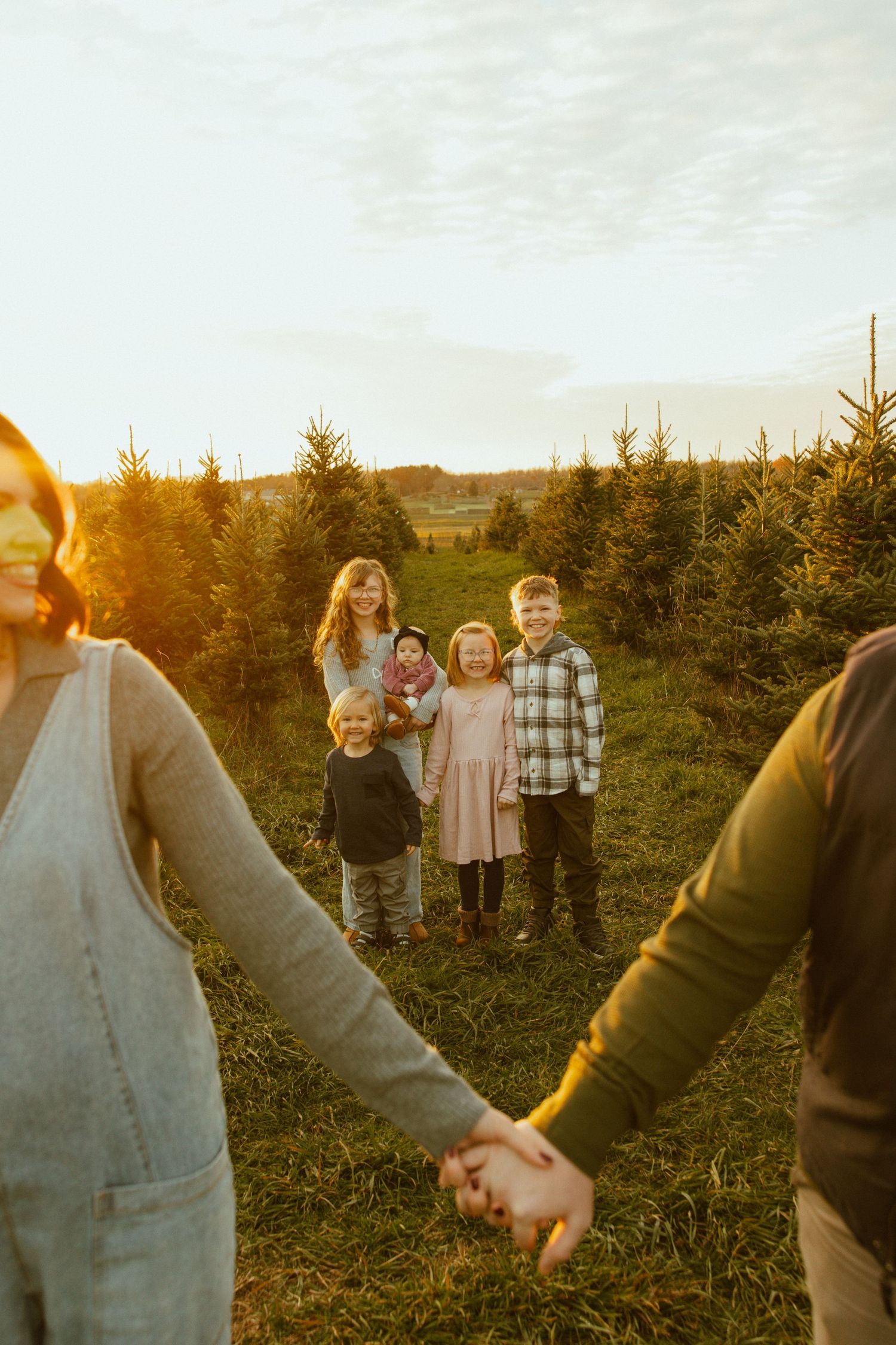 A family poses together among evergreen trees at sunset while two people hold hands in the foreground.