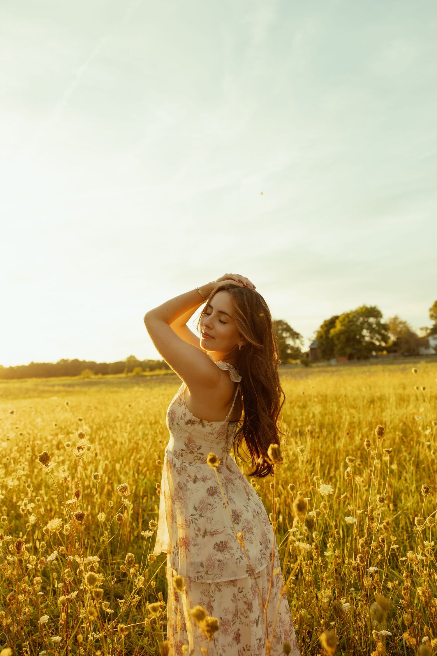 Woman in floral dress poses gracefully in golden wildflower field at sunset with warm summer lighting.