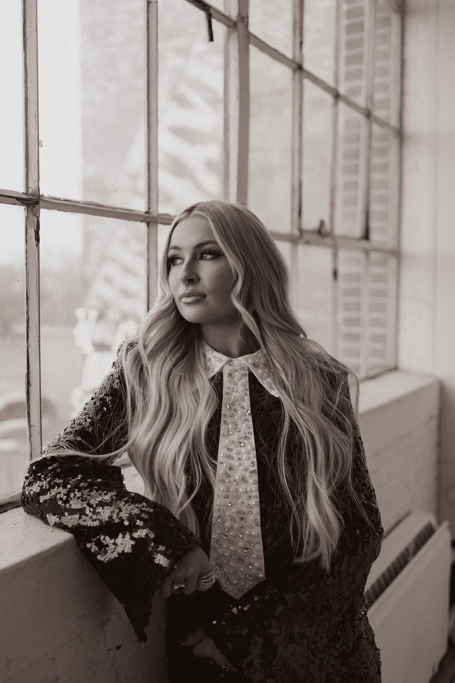 Artistic black and white portrait by window featuring sequined jacket and polka dot tie.