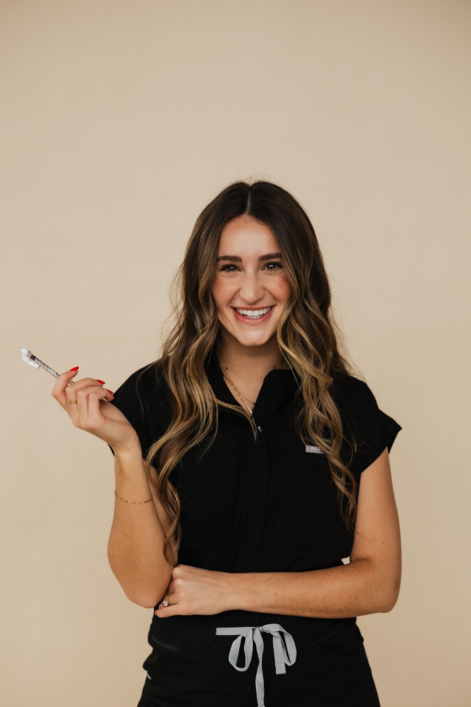 Professional woman in black outfit with long wavy hair smiles while holding pen against beige background.