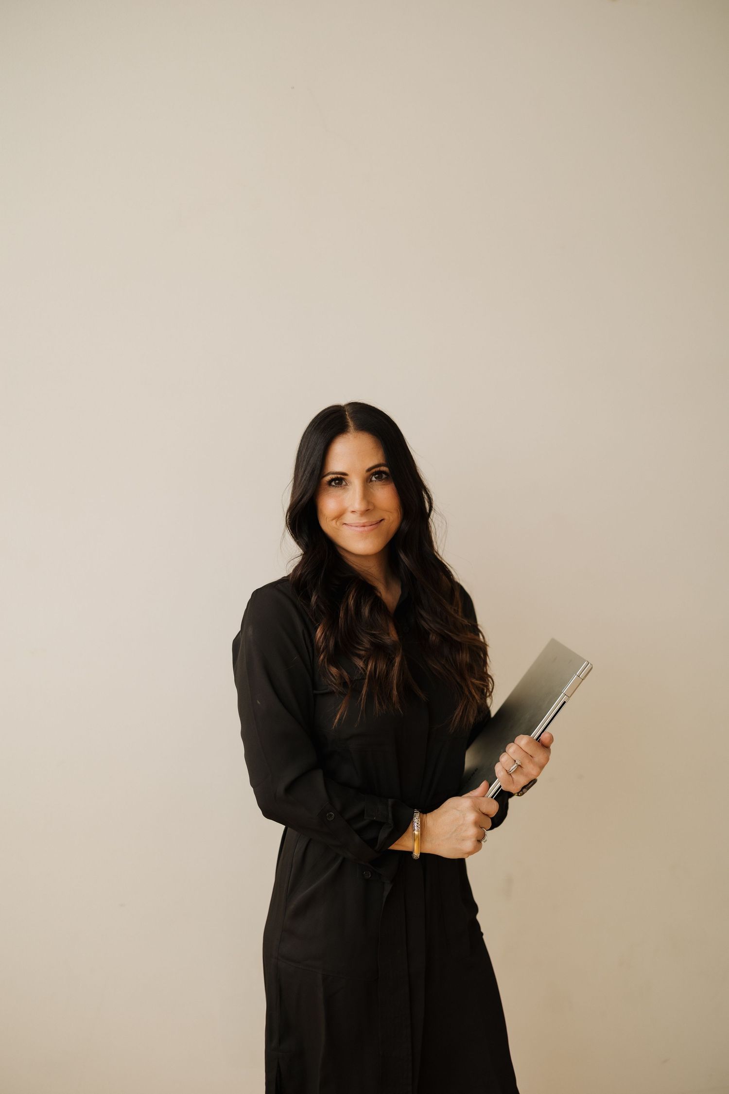 Professional portrait in black outfit holding tablet against neutral background.