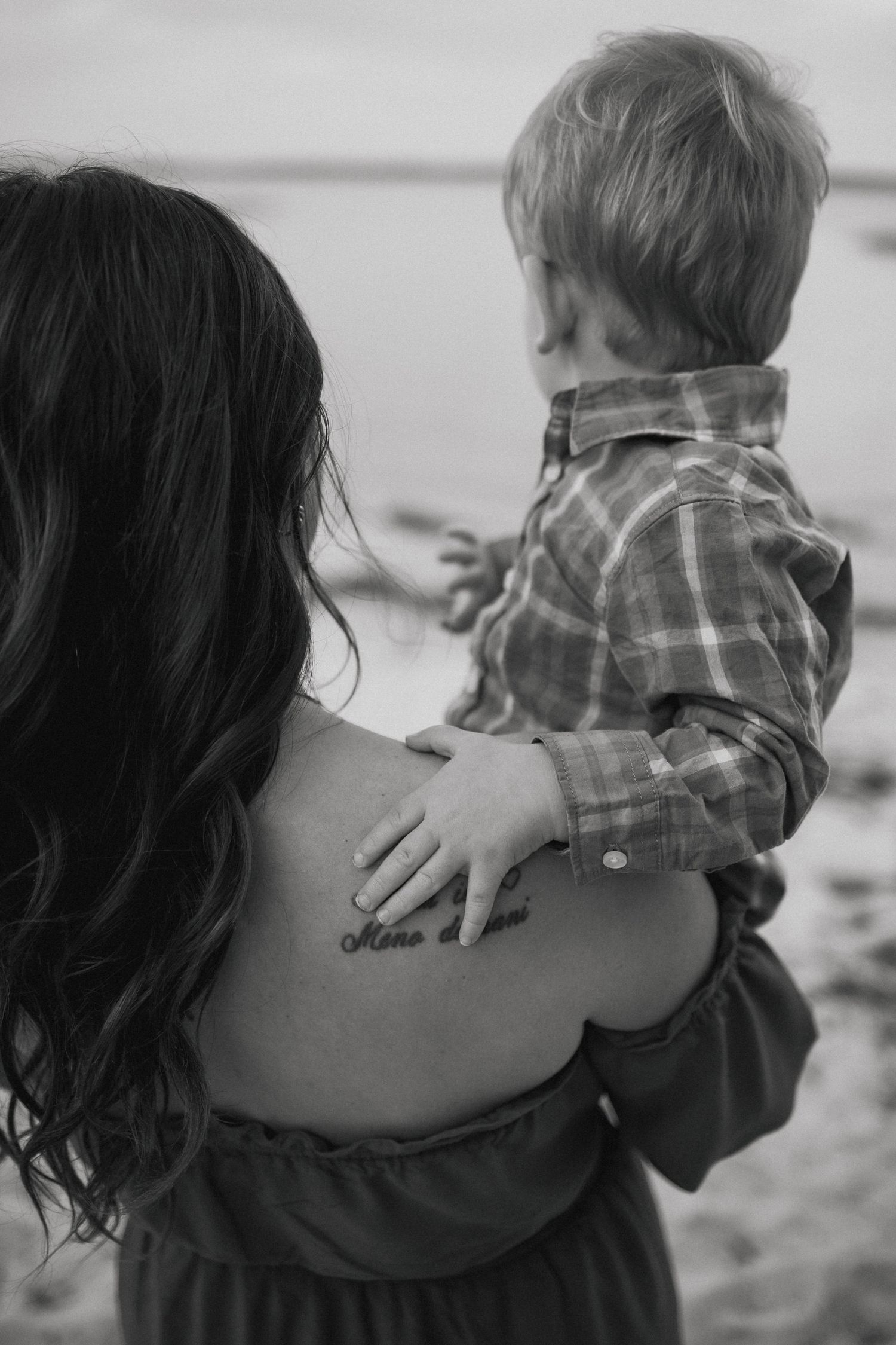 Black and white photo showing child held against parent's back with visible tattoo.