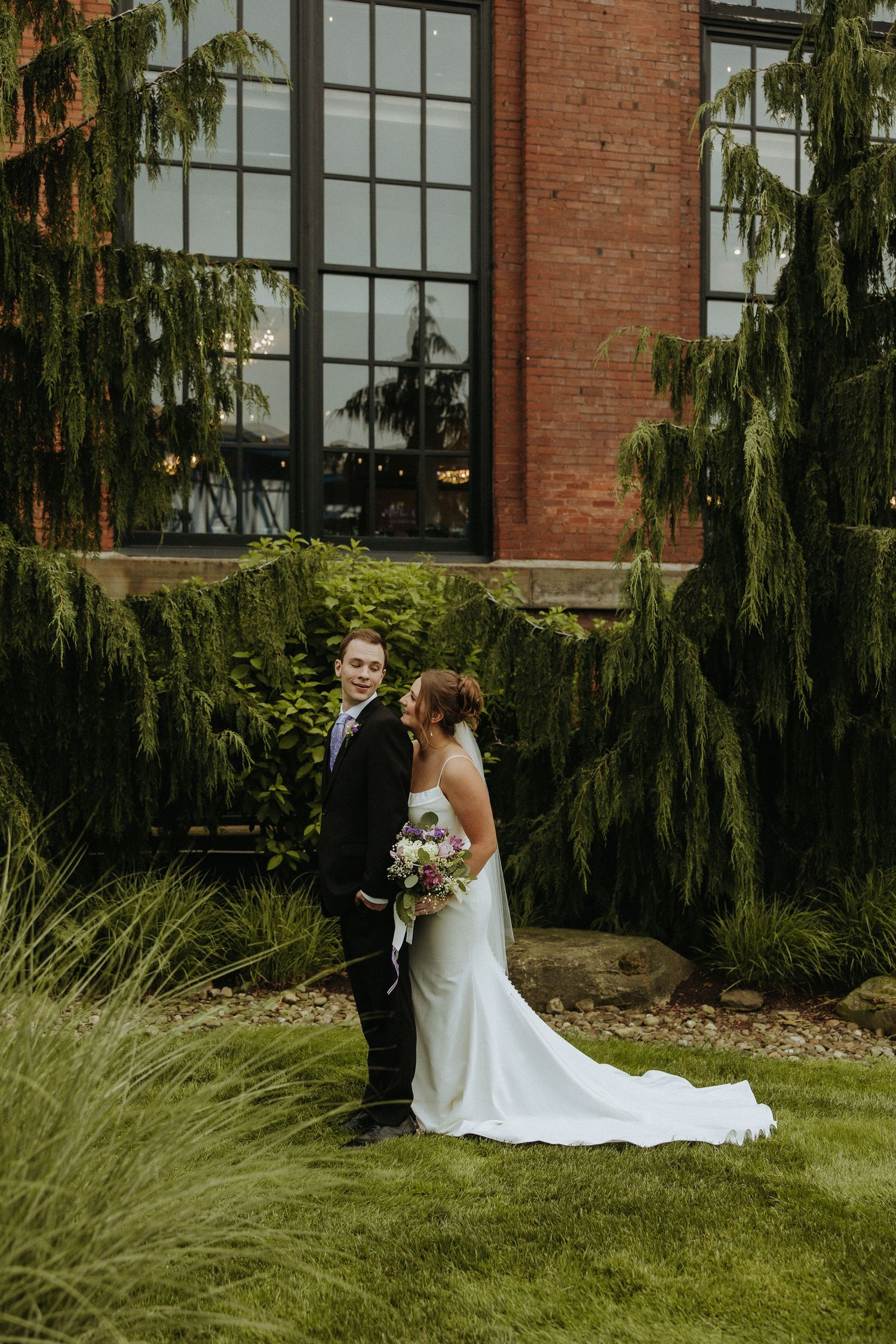 Wedding couple poses in front of a brick building with evergreen trees in an outdoor garden setting.