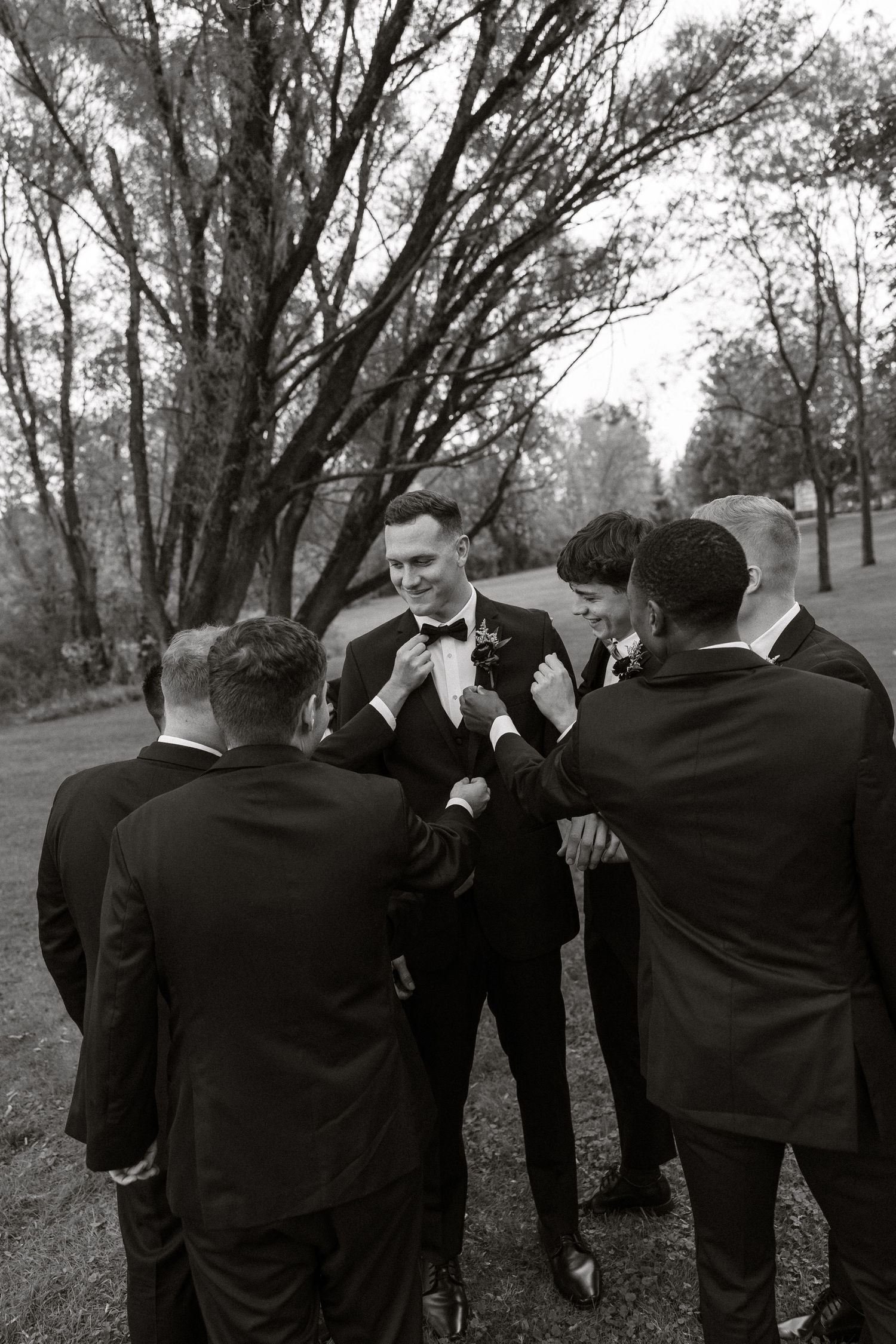 Groomsmen in black suits stand in a line against a backdrop of trees.
