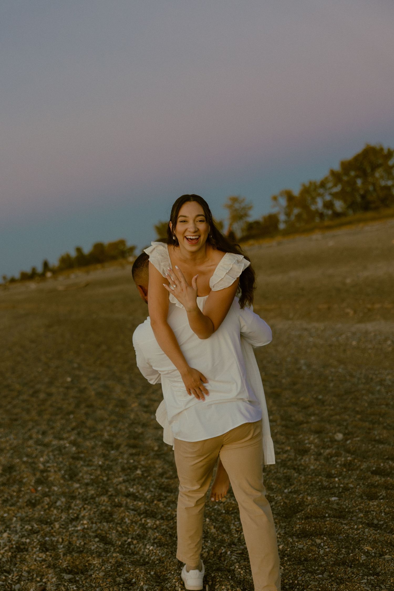 A person in a white top and beige pants poses joyfully in a grassy field at sunset.
