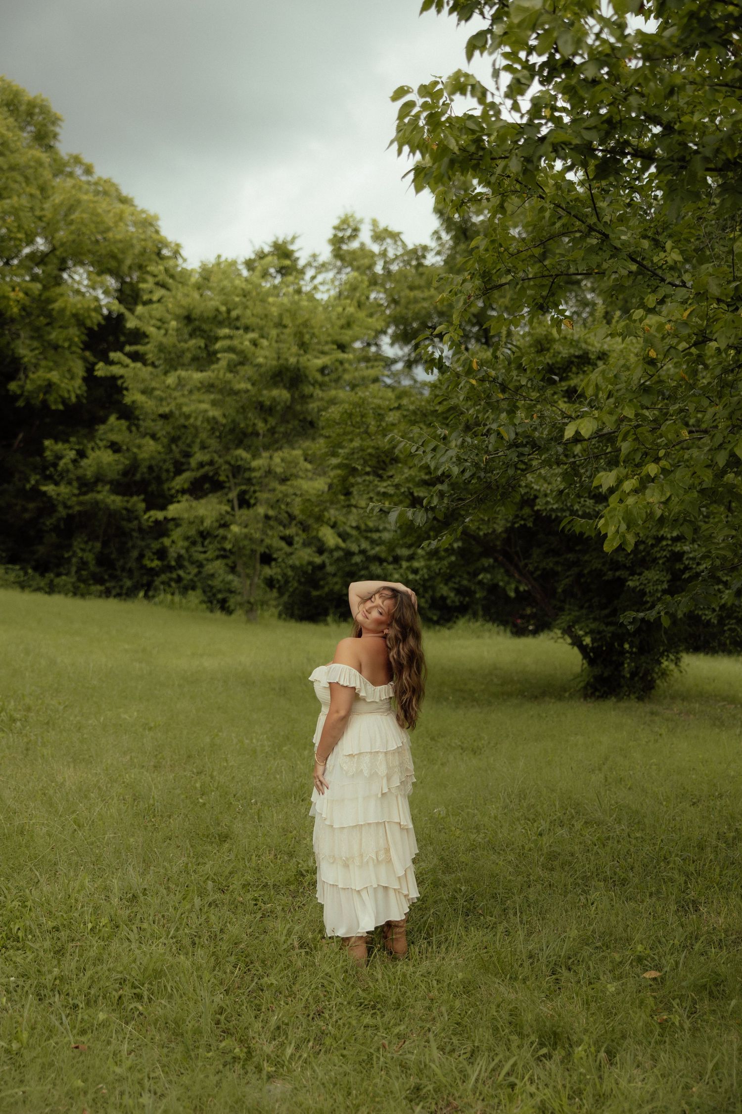 A dreamy outdoor portrait shows a person in a white tiered dress standing in a lush green field surrounded by trees.