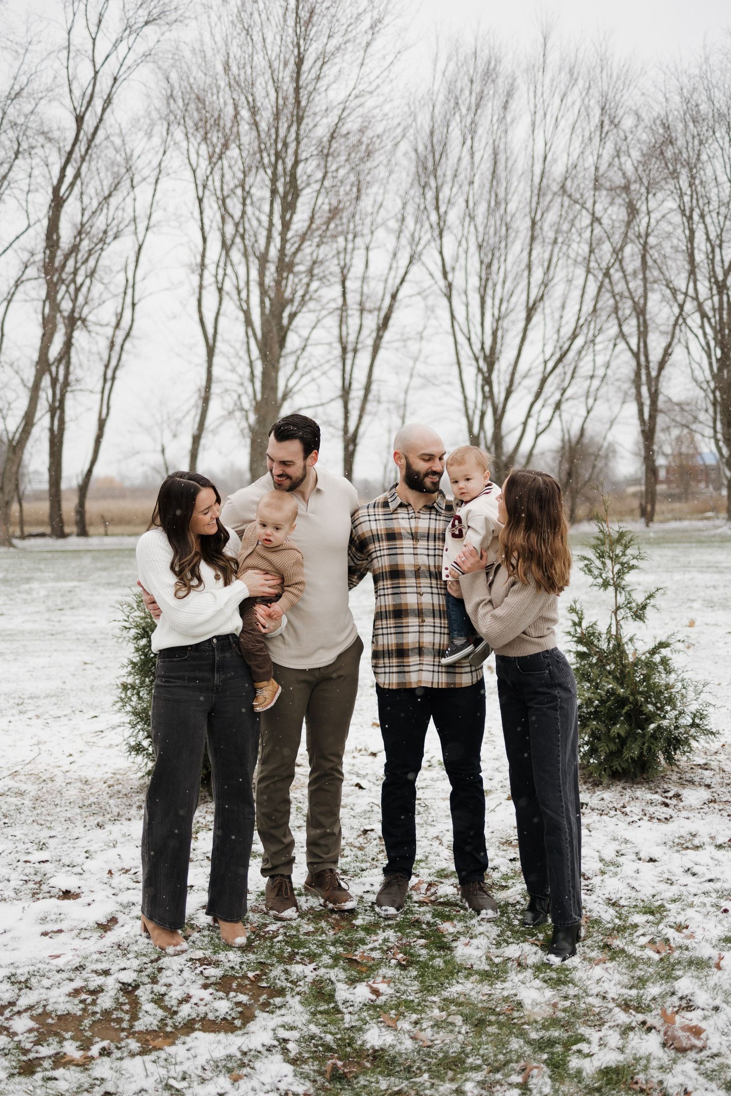 Two couples with children gather in snowy setting near evergreen shrubs.