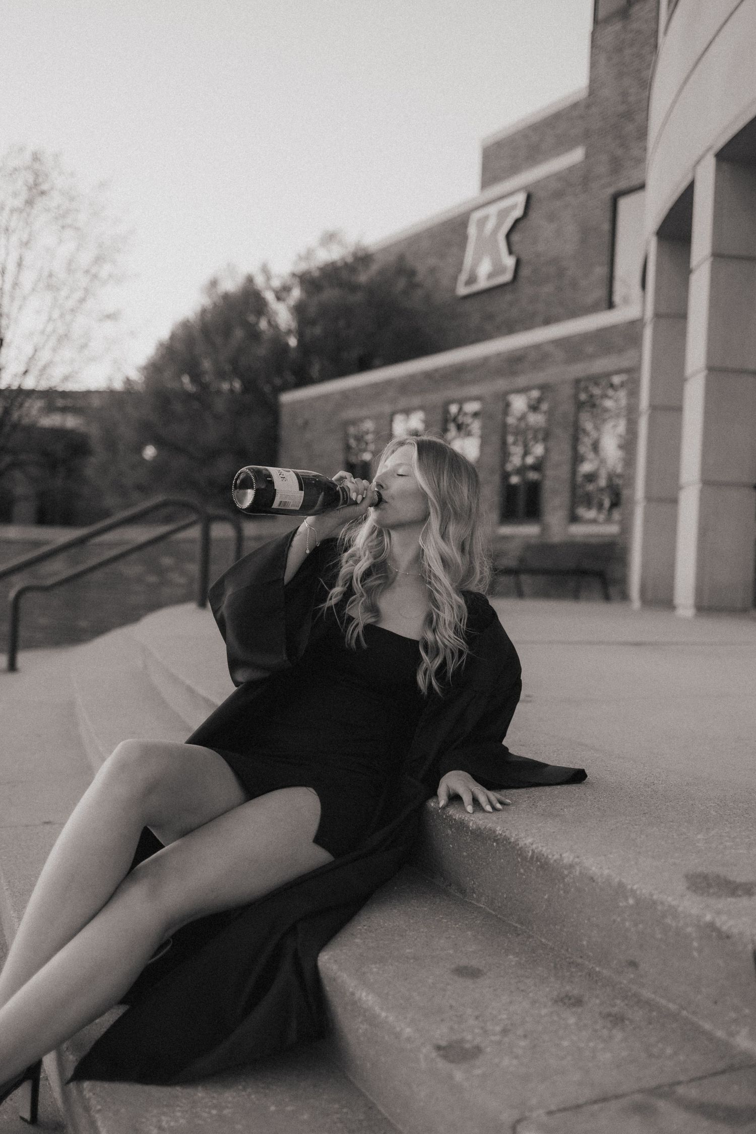 A graduate in black attire poses dramatically on steps while drinking from graduation cap.