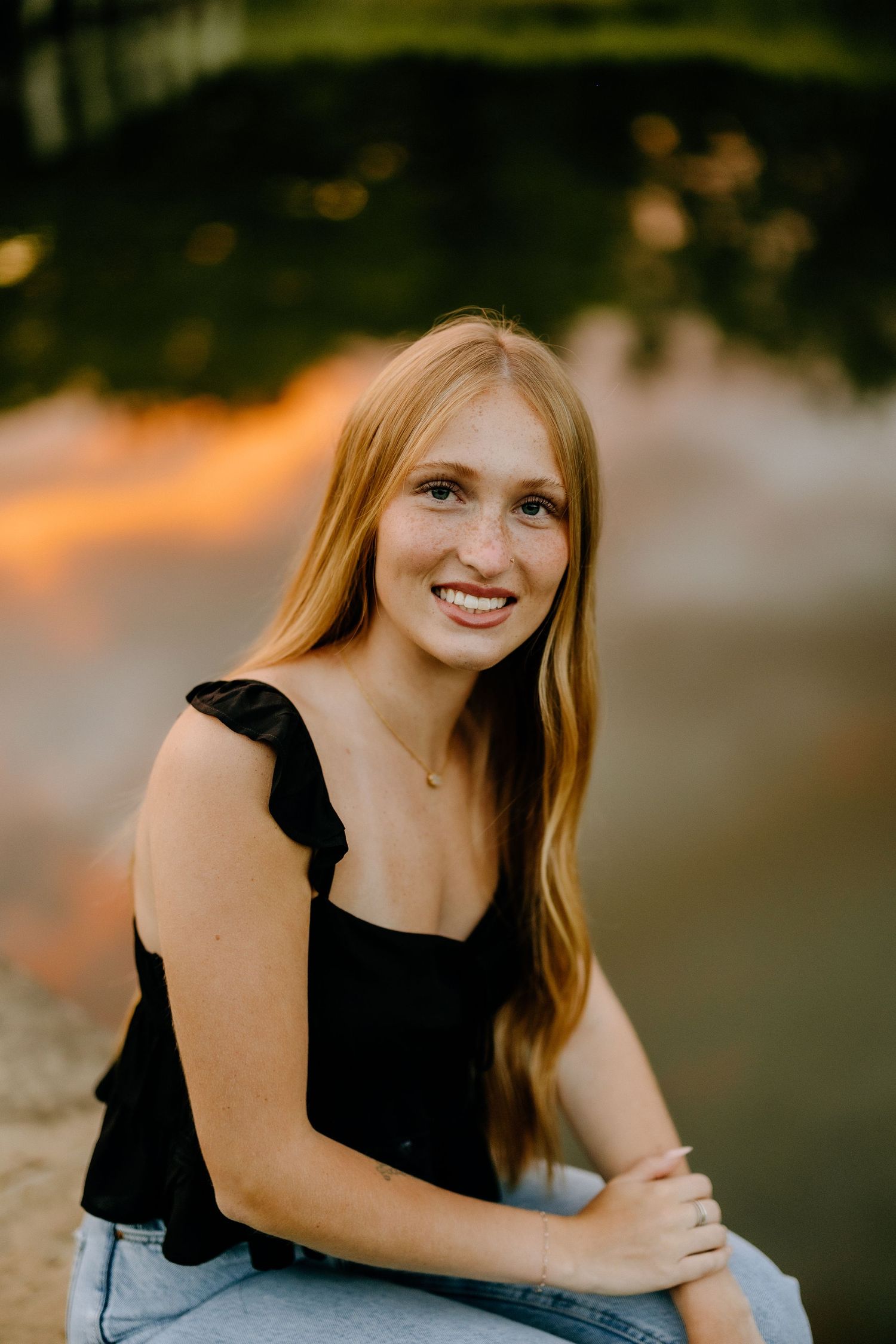 A person in a black sleeveless top and jeans smiles during golden hour outdoor portrait session.