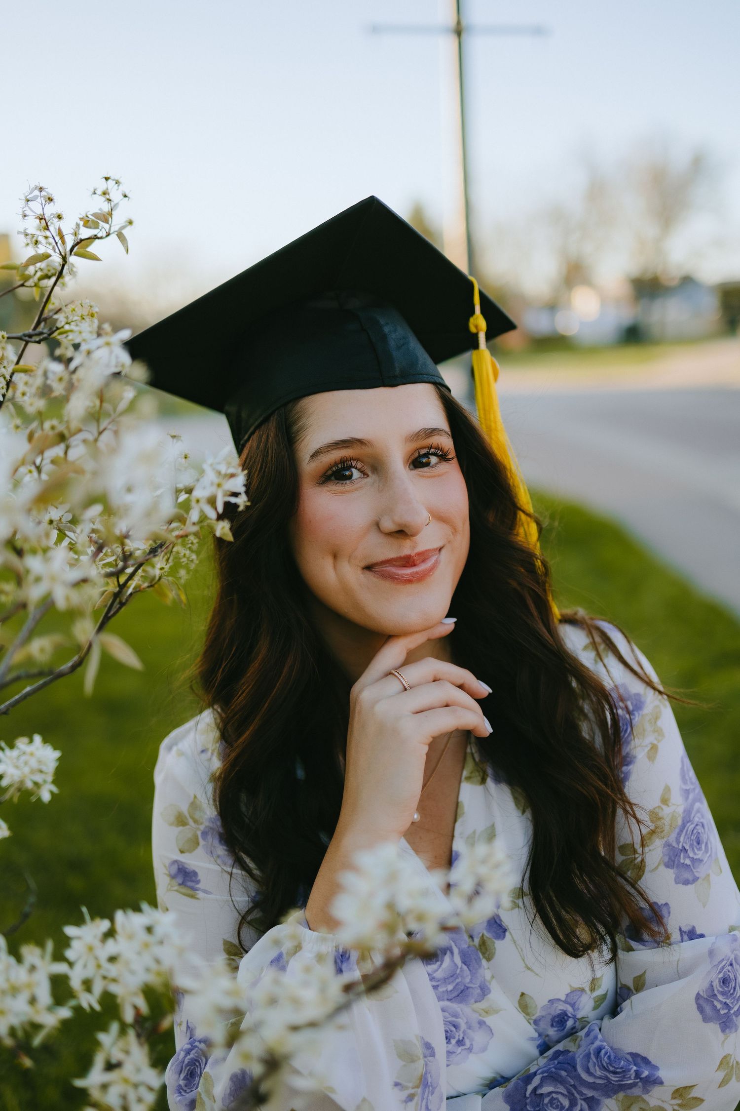A graduate in a black cap and floral dress poses thoughtfully outdoors among white flowers.