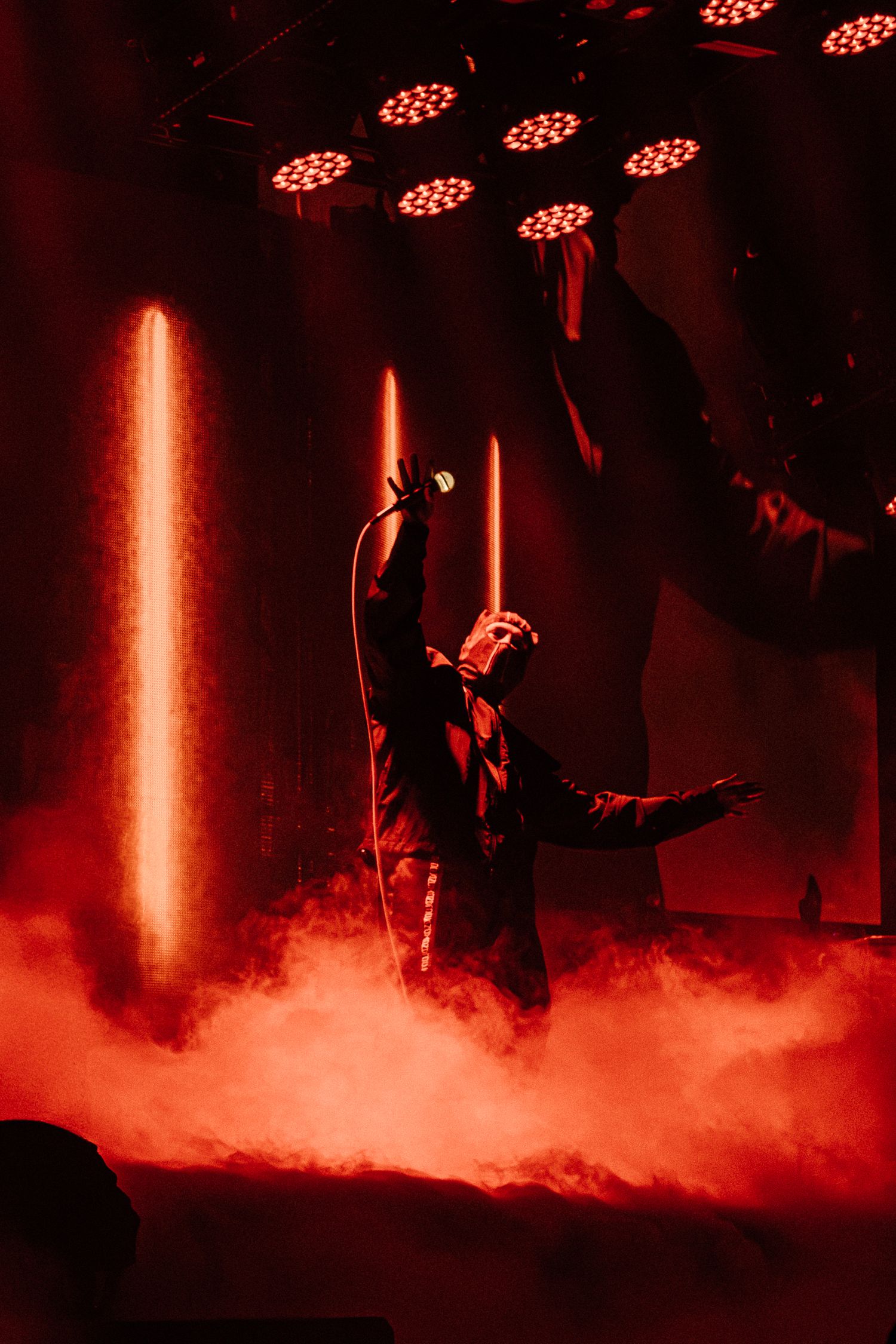 Concert performer in dramatic pose surrounded by red stage lights and atmospheric smoke.