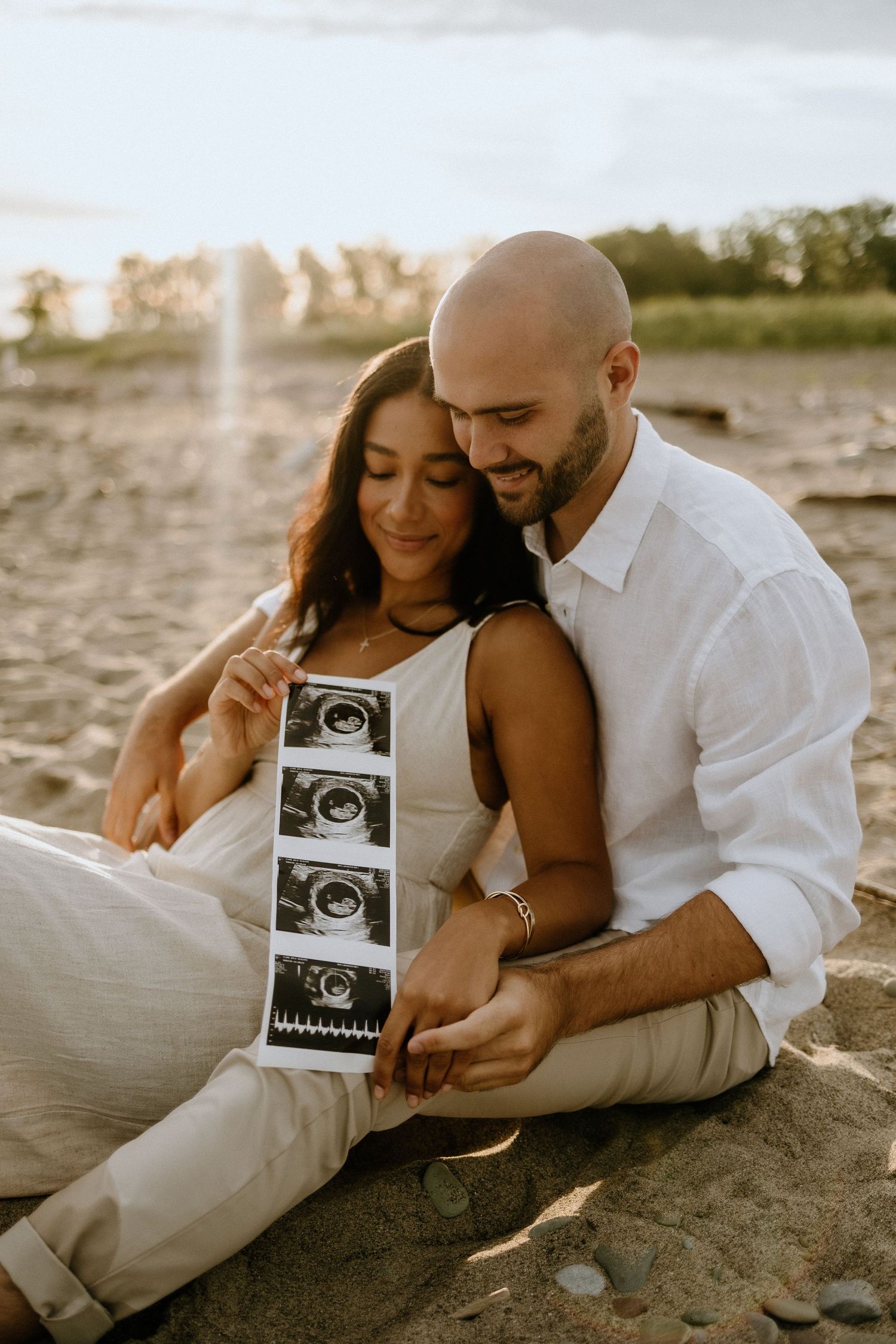 Expecting couple sits on beach sand while holding ultrasound images during pregnancy announcement photoshoot.