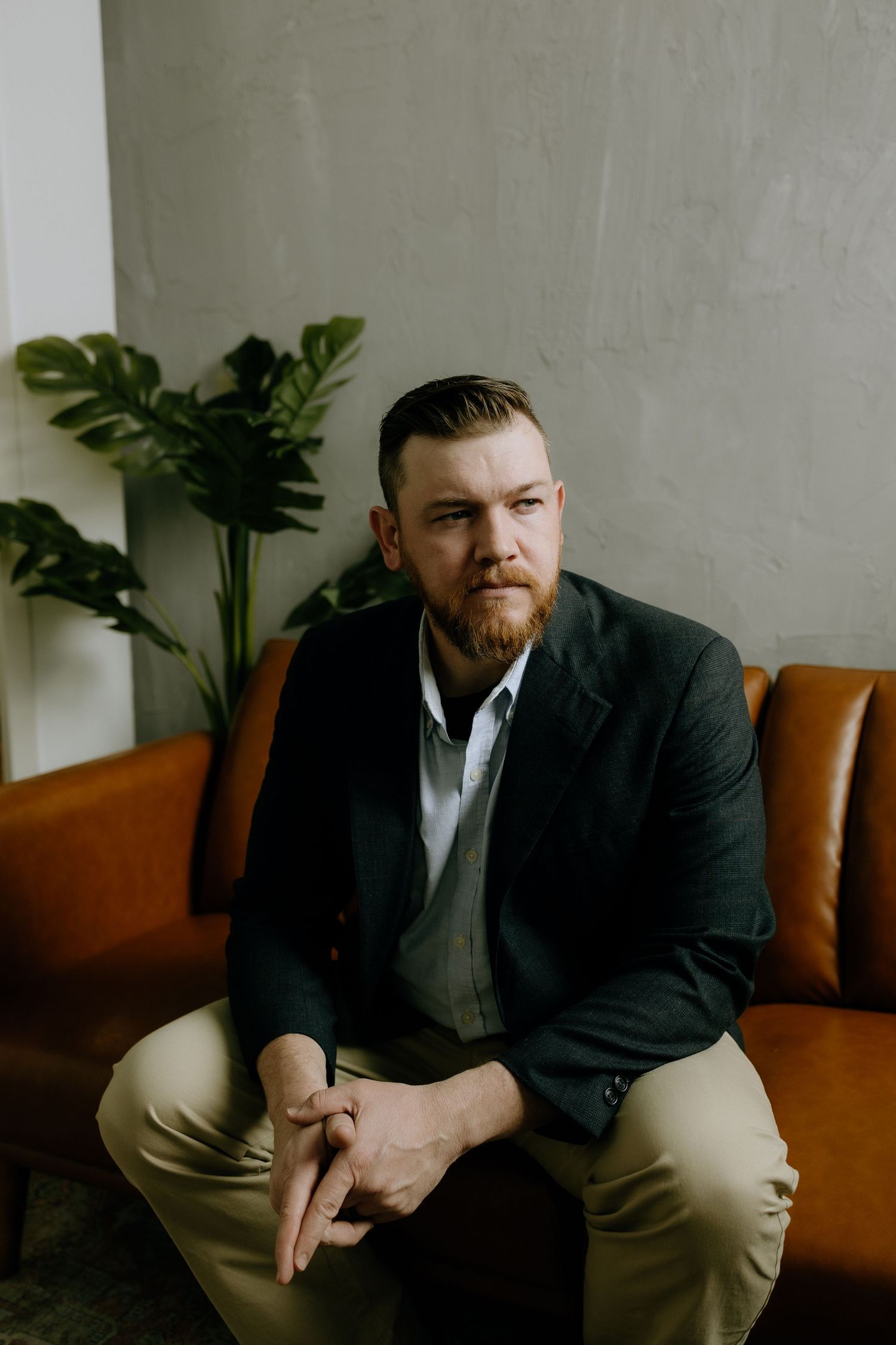 Professional portrait in dark suit seated on brown leather couch with plants in background.