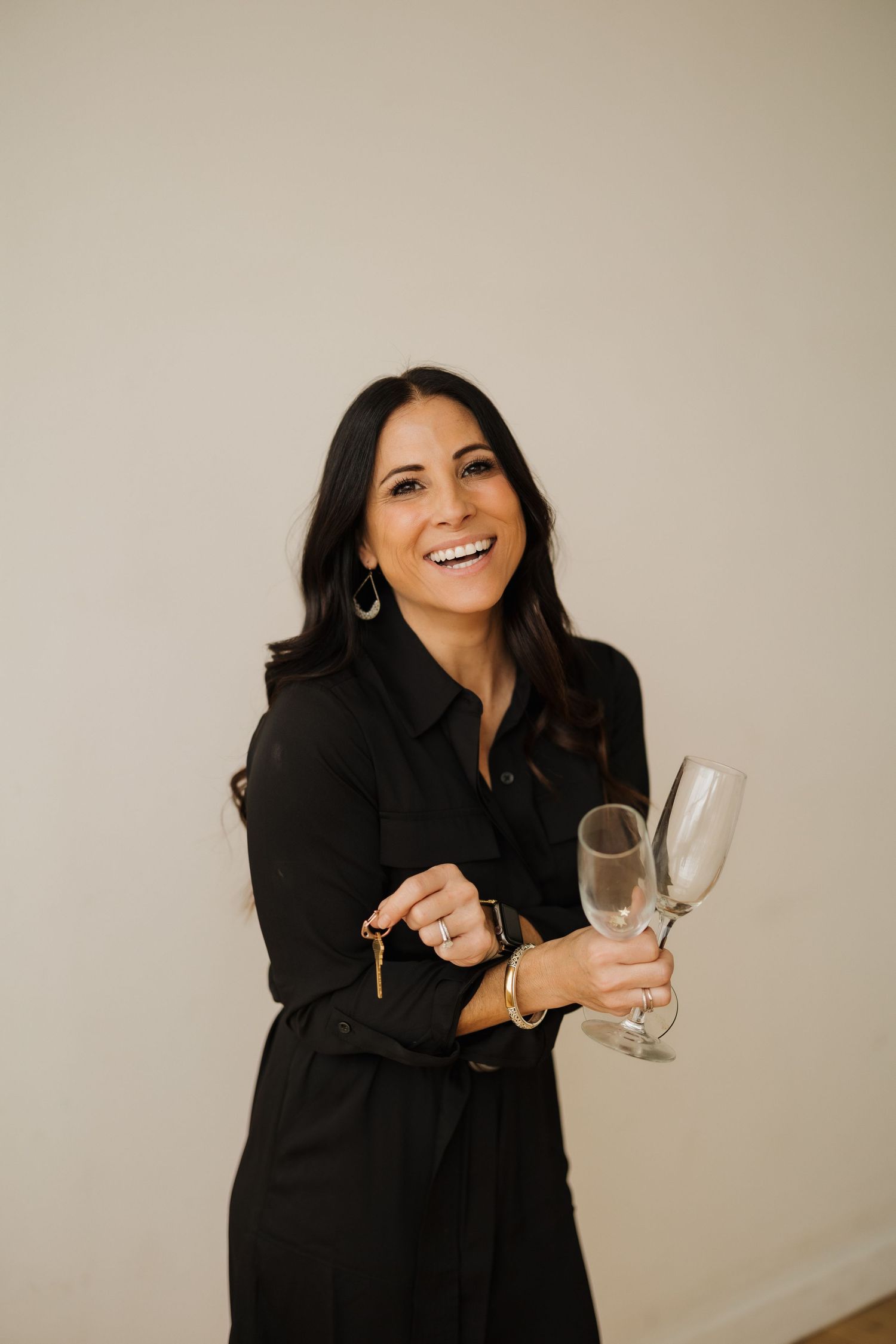Professional wearing a black dress holds champagne glasses while smiling against neutral wall.