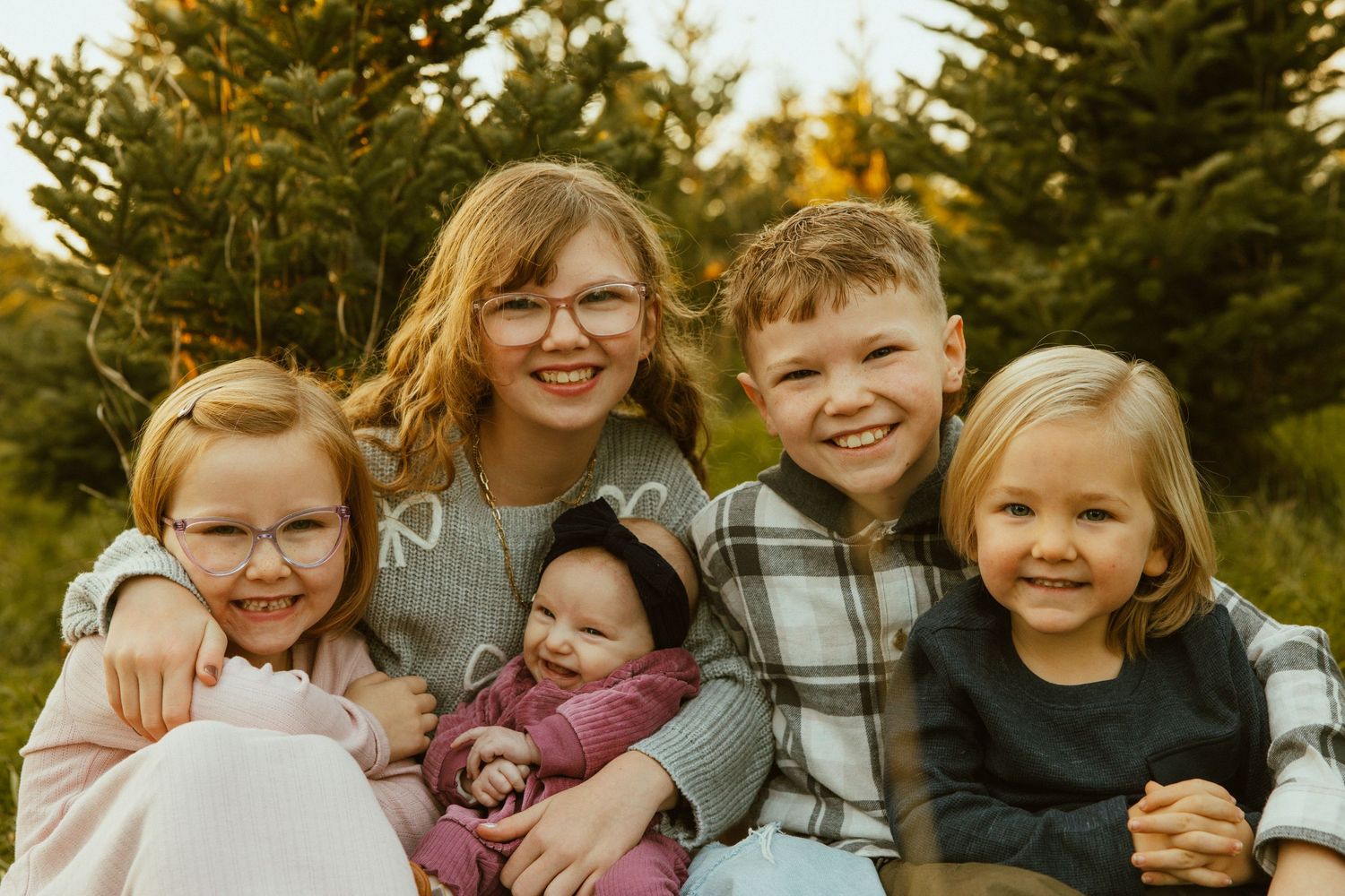 A group of children sitting together outdoors among evergreen trees in warm autumn sunlight.