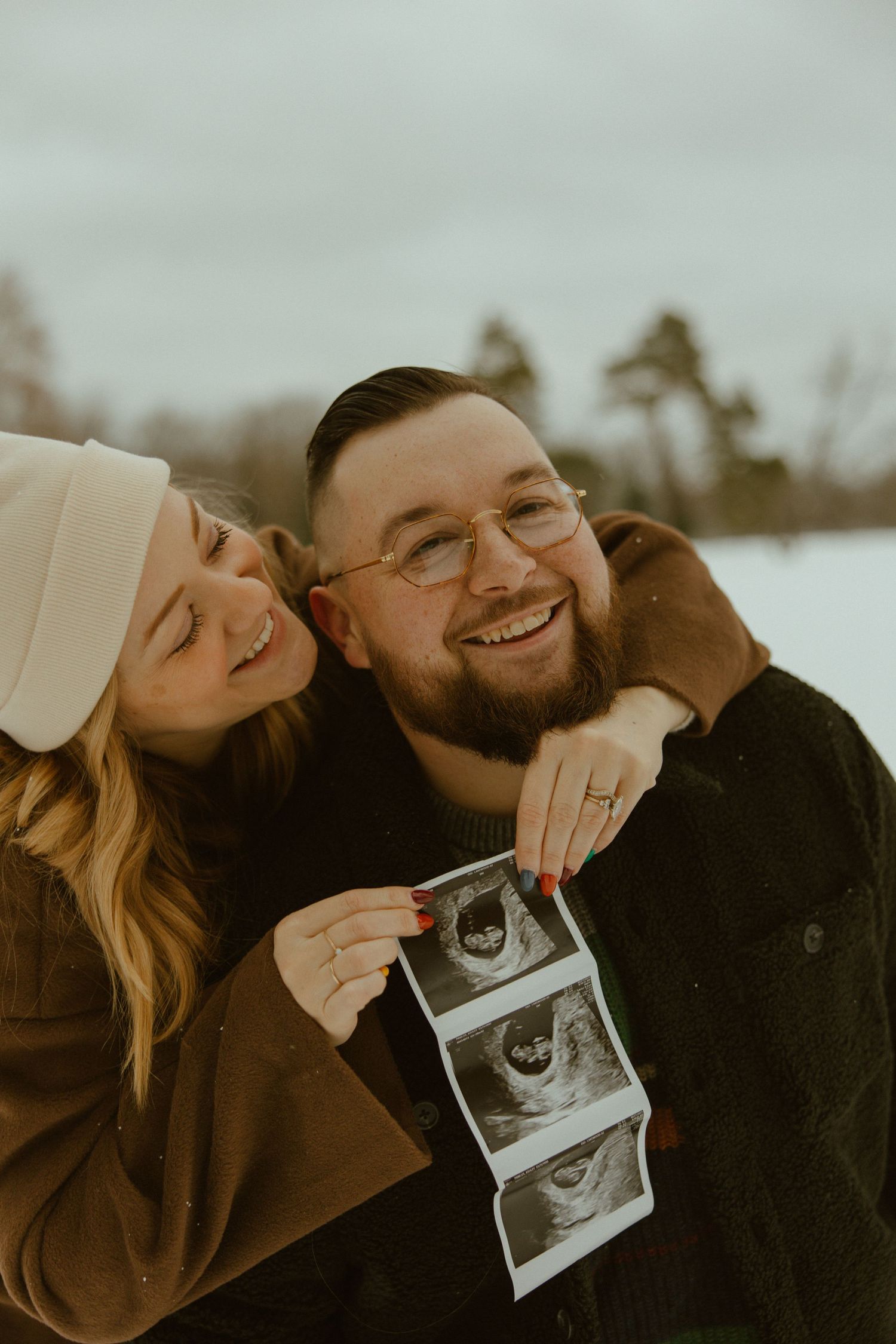 A couple holds up an ultrasound photo while sharing a tender moment together outdoors in muted tones.