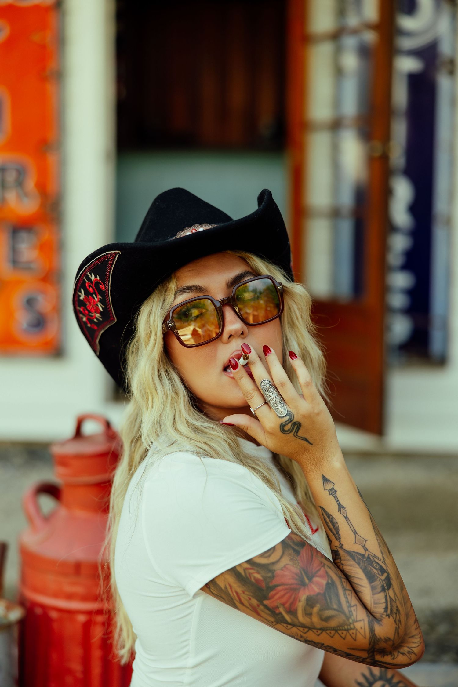 Woman in white t-shirt with tattoos and sunglasses poses next to red post box in urban setting.