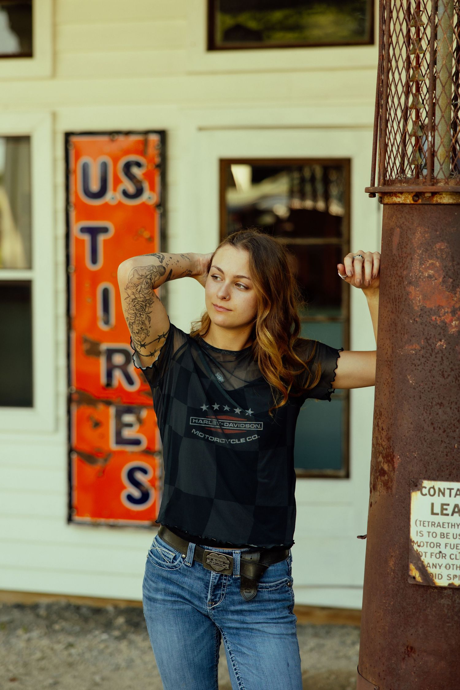 Person in black sweater and jeans leans against rustic metal pole with vintage signs in background.