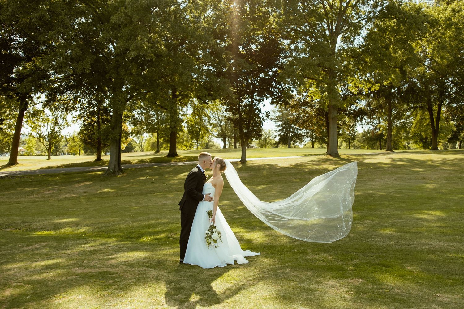 A wedding couple poses in a sunlit park with a flowing veil creating romantic movement in the breeze.