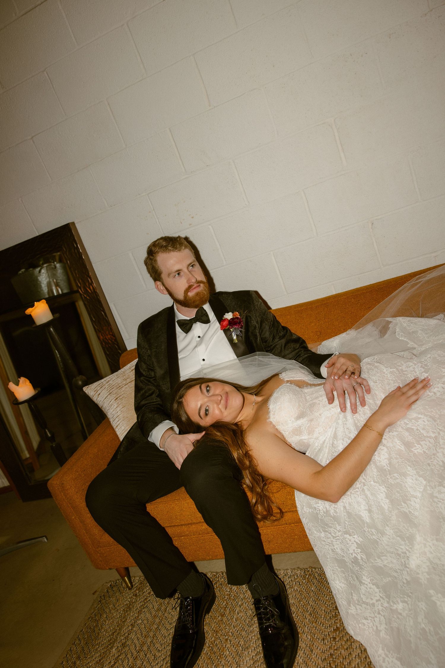 A vintage-style wedding photo of a couple lounging on an orange couch in formal attire.