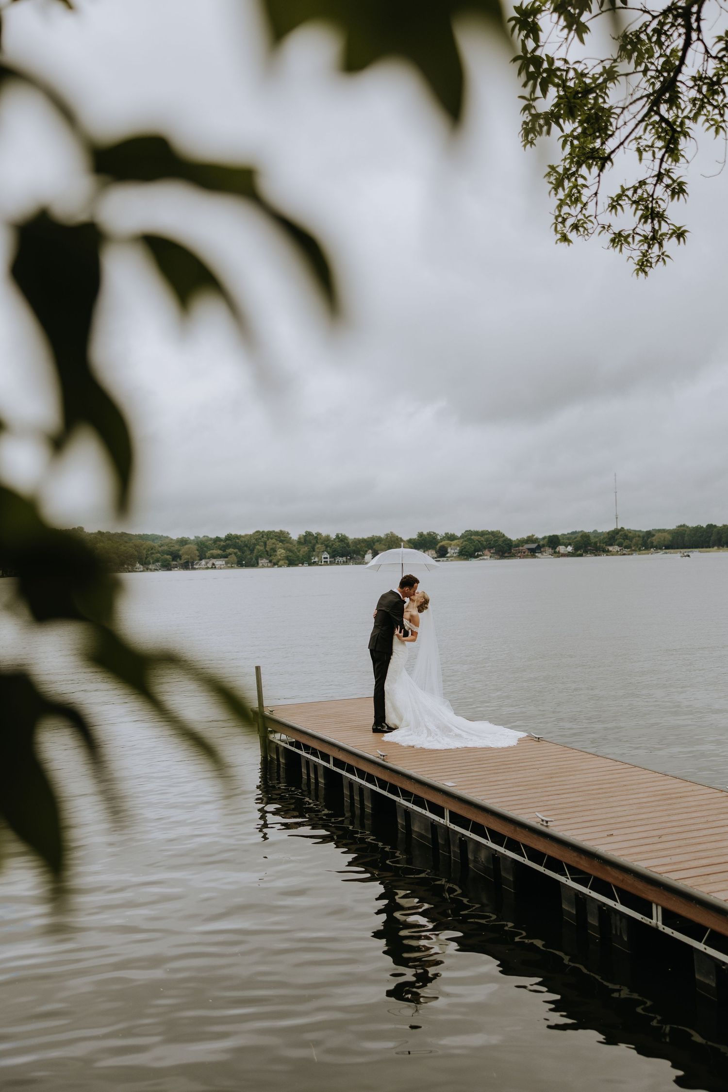 Wedding couple standing on a wooden dock overlooking a misty lake with tree branches framing the scene.