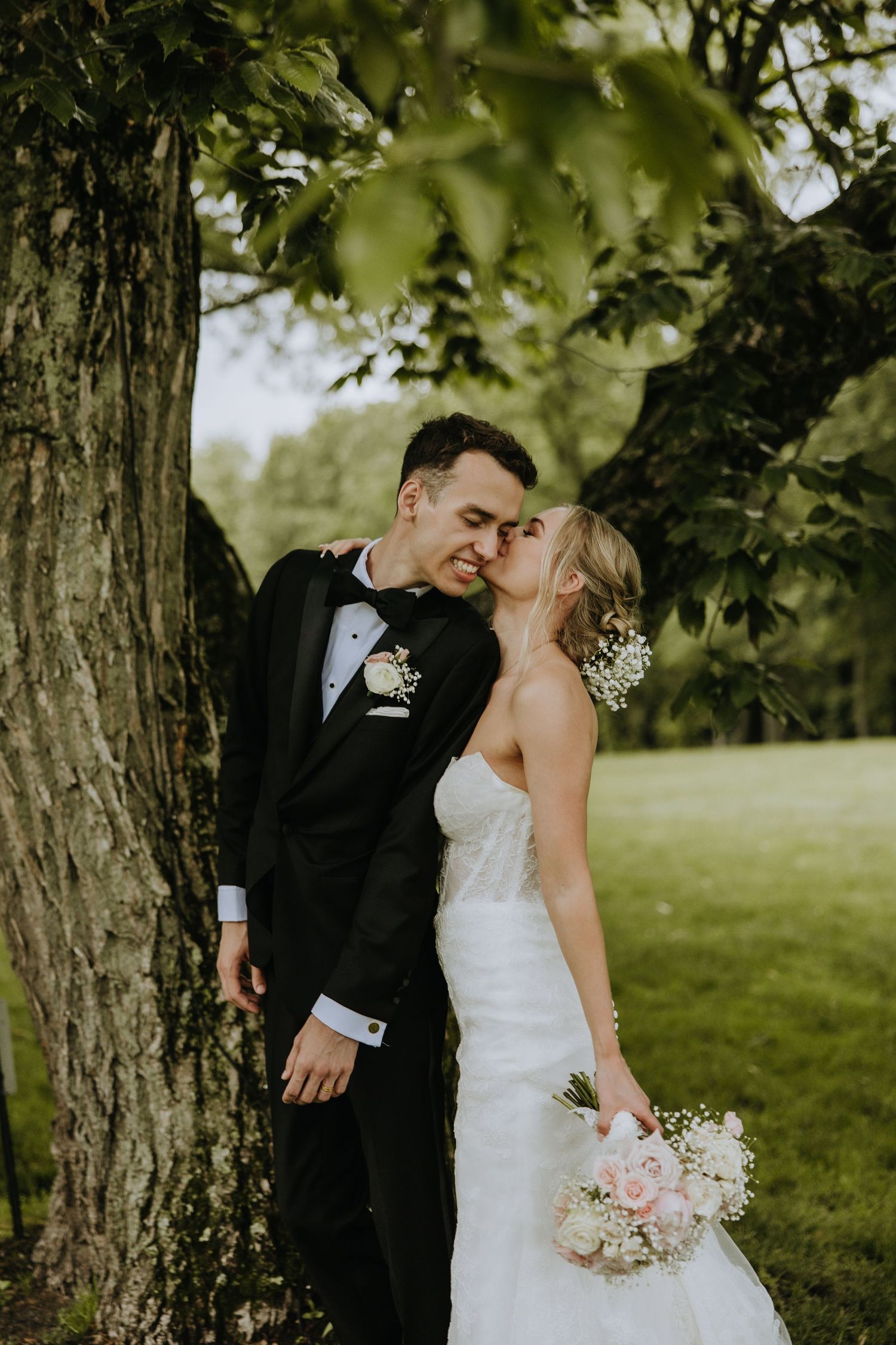A romantic wedding couple shares a tender moment under a large tree in a garden setting with a soft pink bouquet.