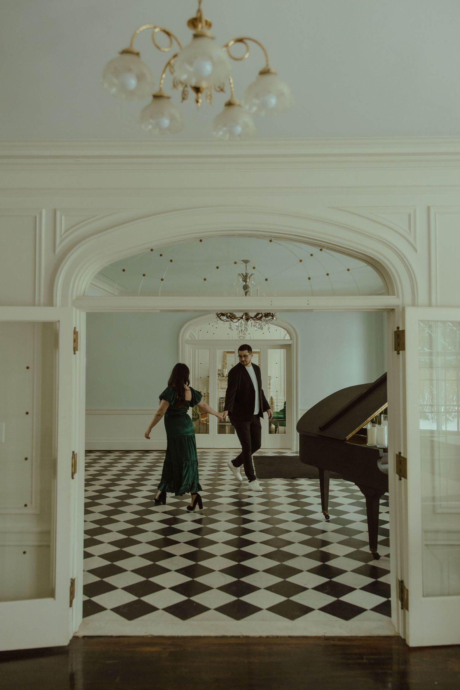 Couples dance in an elegant ballroom with black and white checkered floors and a grand piano under crystal chandeliers.