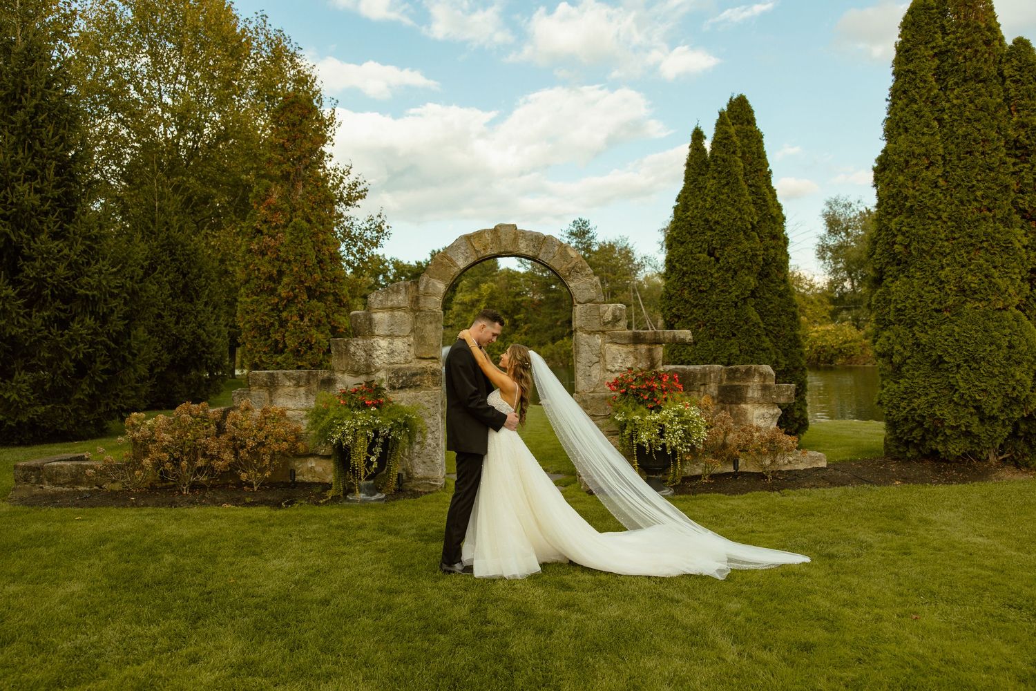 A couple embraces in front of a stone archway in a garden setting with tall cypress trees and blue sky.