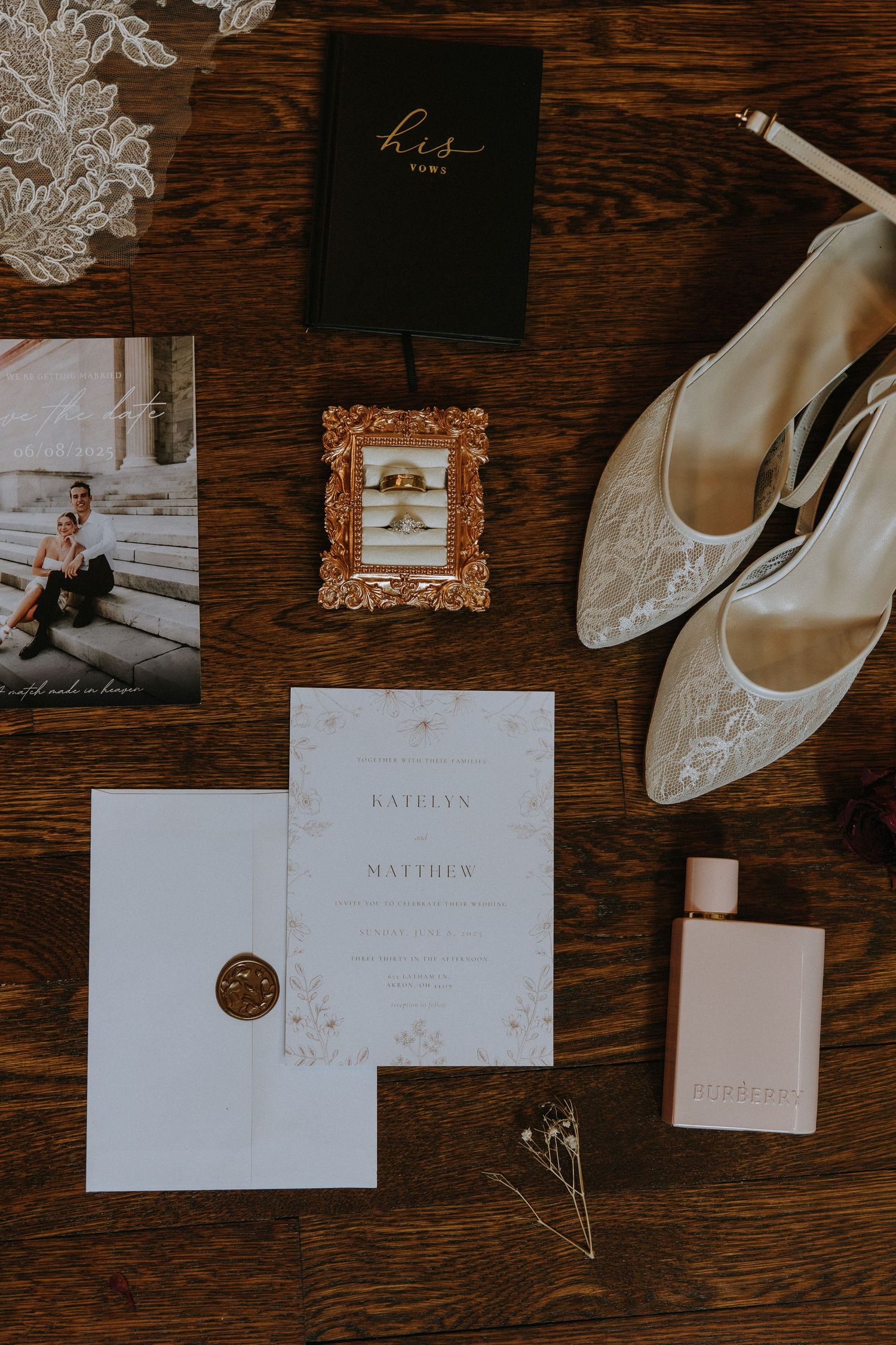 Wedding details flatlay with invitation, white lace shoes, perfume bottle and vintage frame on dark wooden surface.